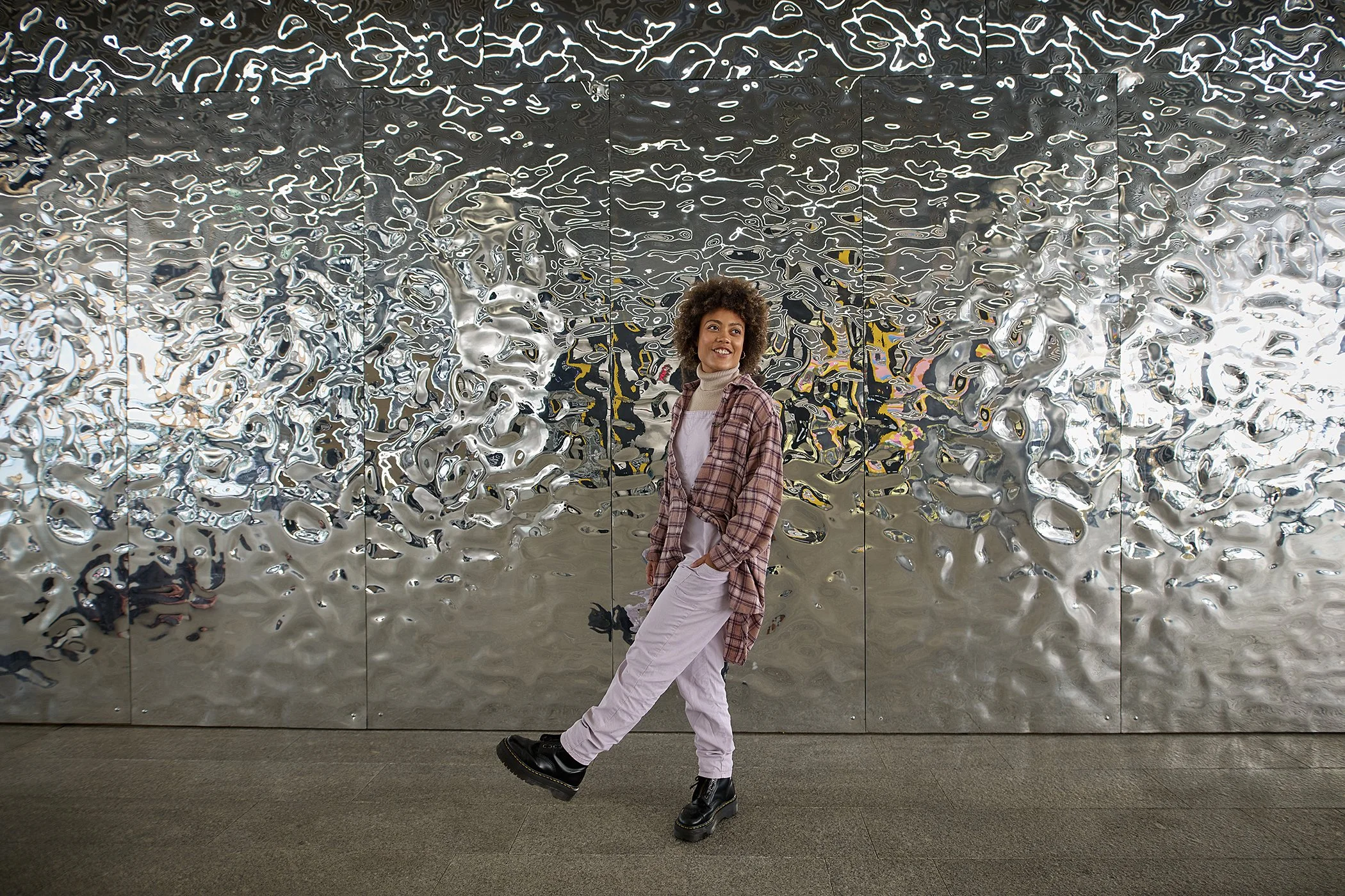 A woman with curly hair smiling, wearing a plaid shirt, white pants, and black boots, standing against a reflective, metallic, textured wall.