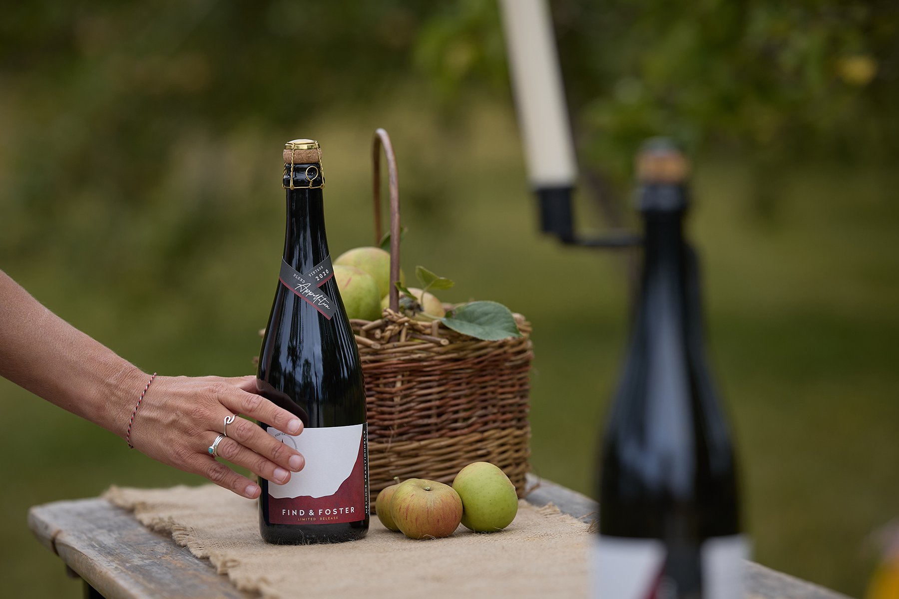 A hand holding a bottle of Find & Foster fine cider against a blurred Devon orchard backdrop.