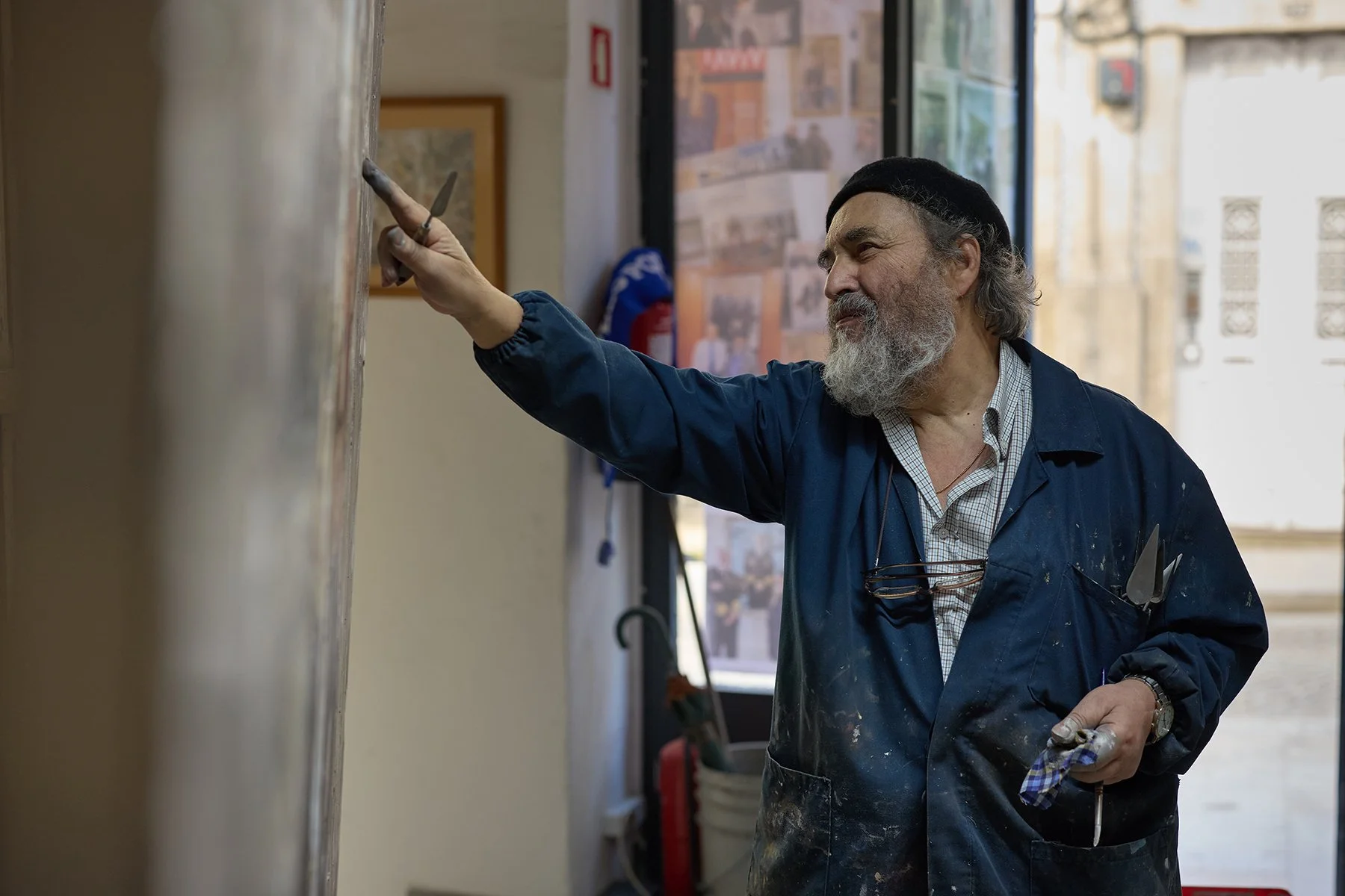 An older man with a gray beard is painting on a wall in an art studio, wearing a black beret and a blue painter's smock.