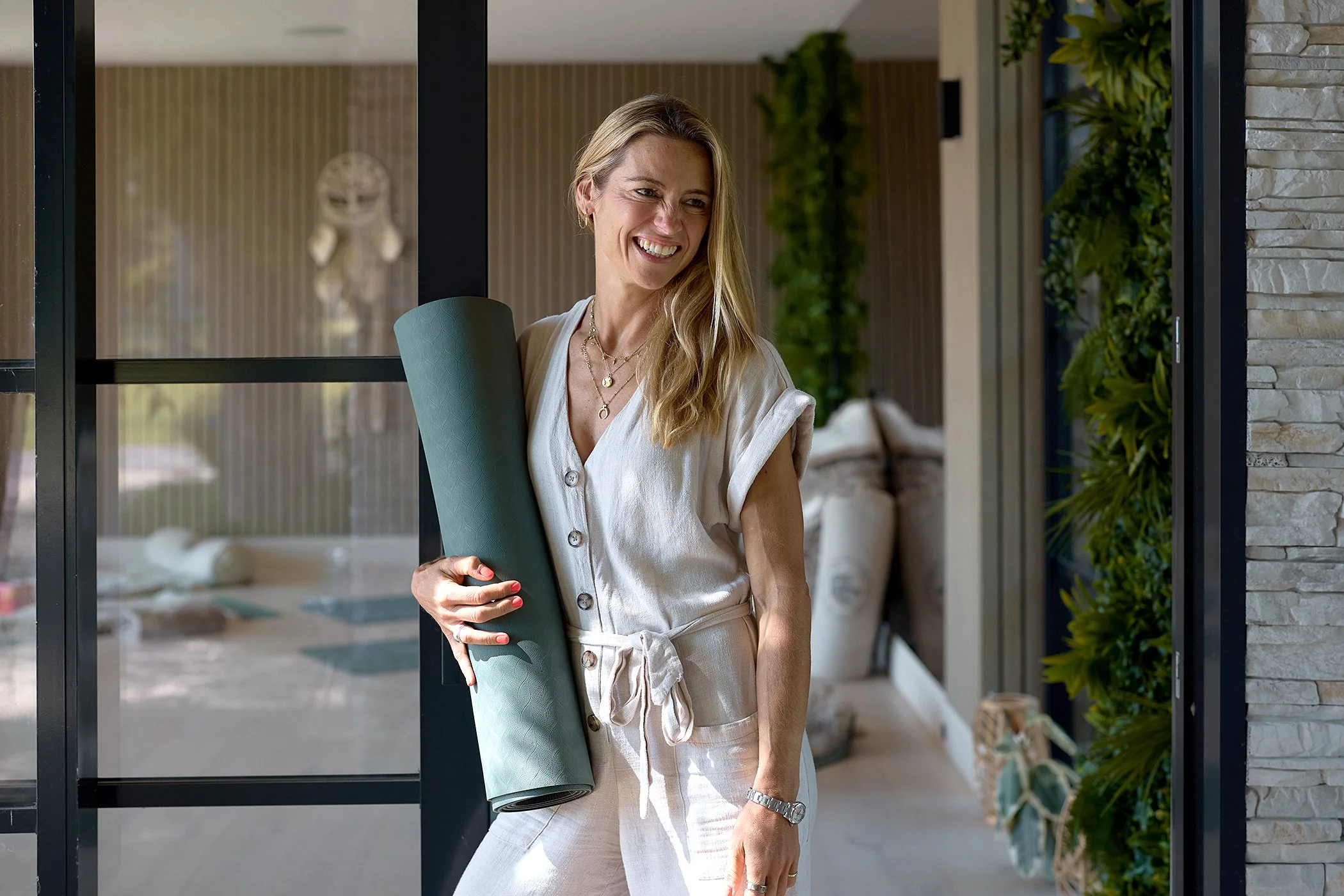A woman holding a yoga mat and smiling inside a modern home or yoga studio, with green plants and stone wall decor in the background.