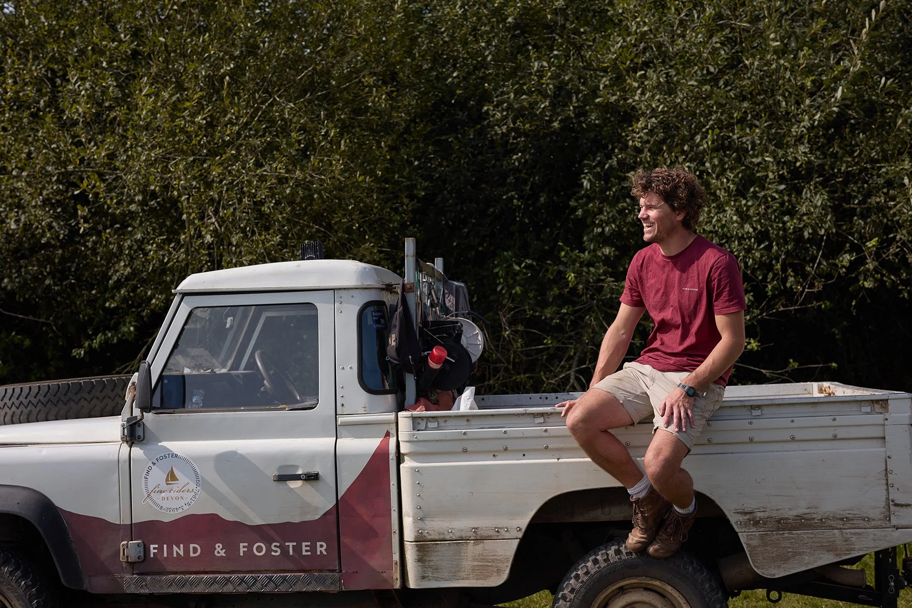 A man sitting on the back of a white Find & Foster pickup truck, smiling, with greenery in the background.