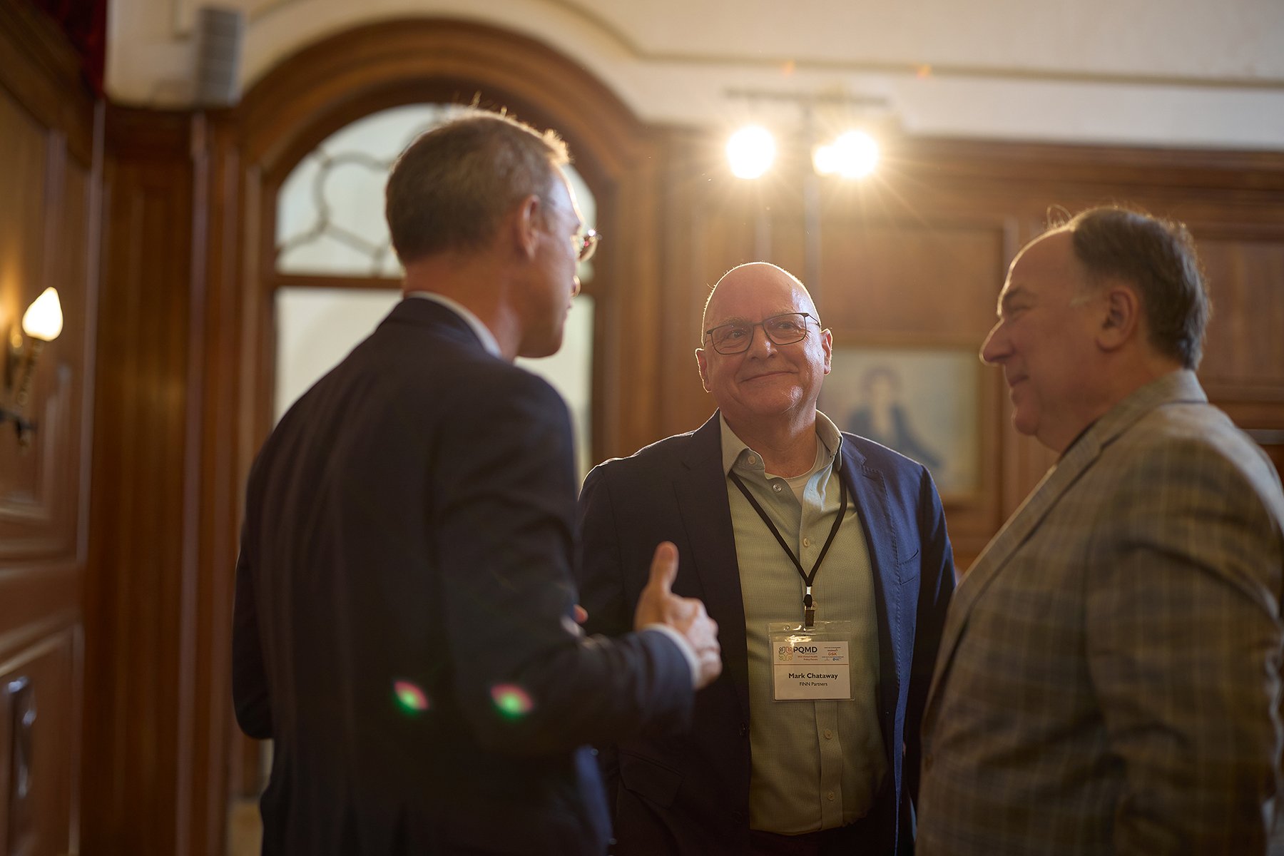 Three men engaged in conversation at a formal event in a wood-paneled room with a portrait painting in the background, lights shining overhead.