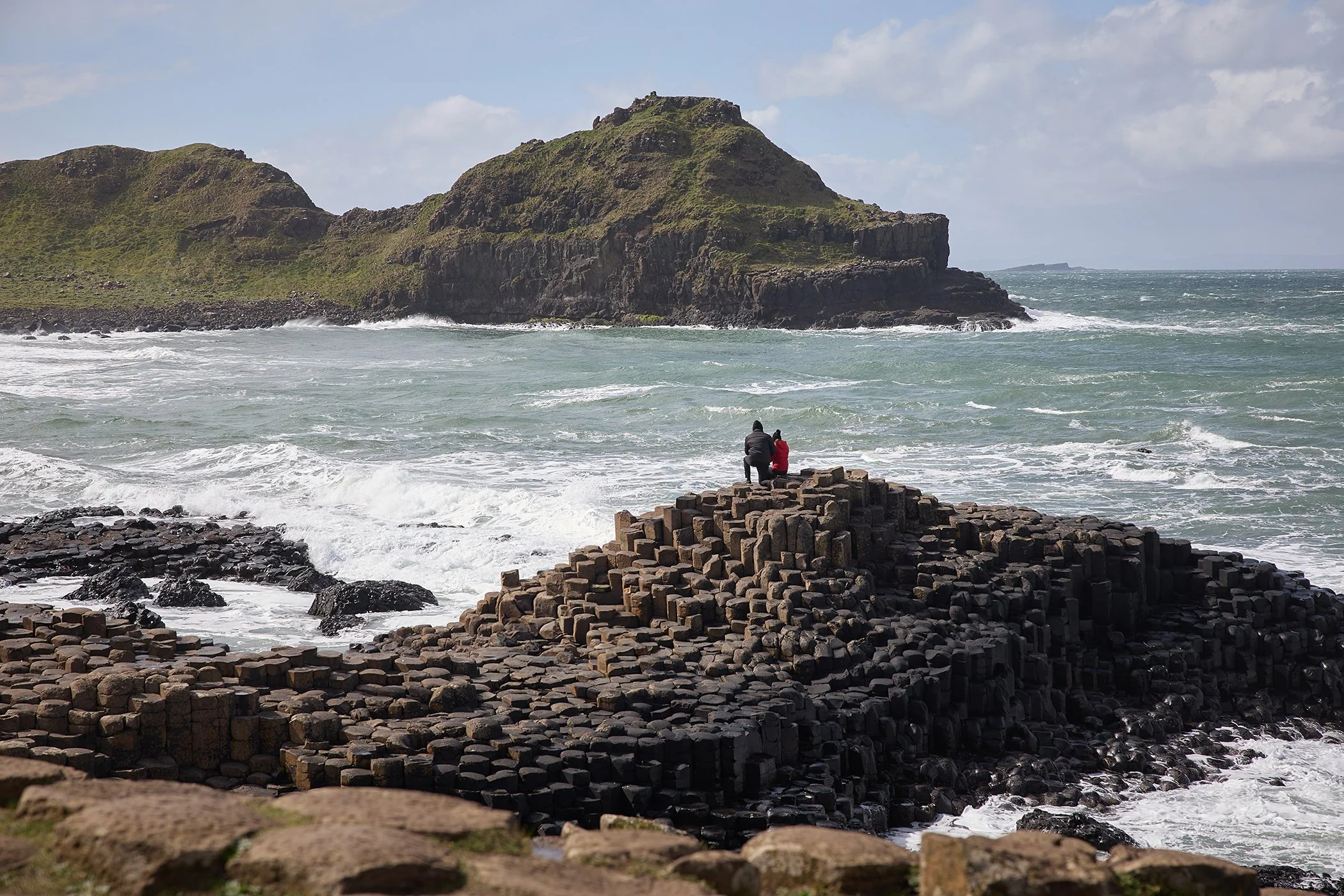 Two people sitting on basalt columns on a rocky shoreline, overlooking the ocean with waves crashing and a green, rocky hill in the background.