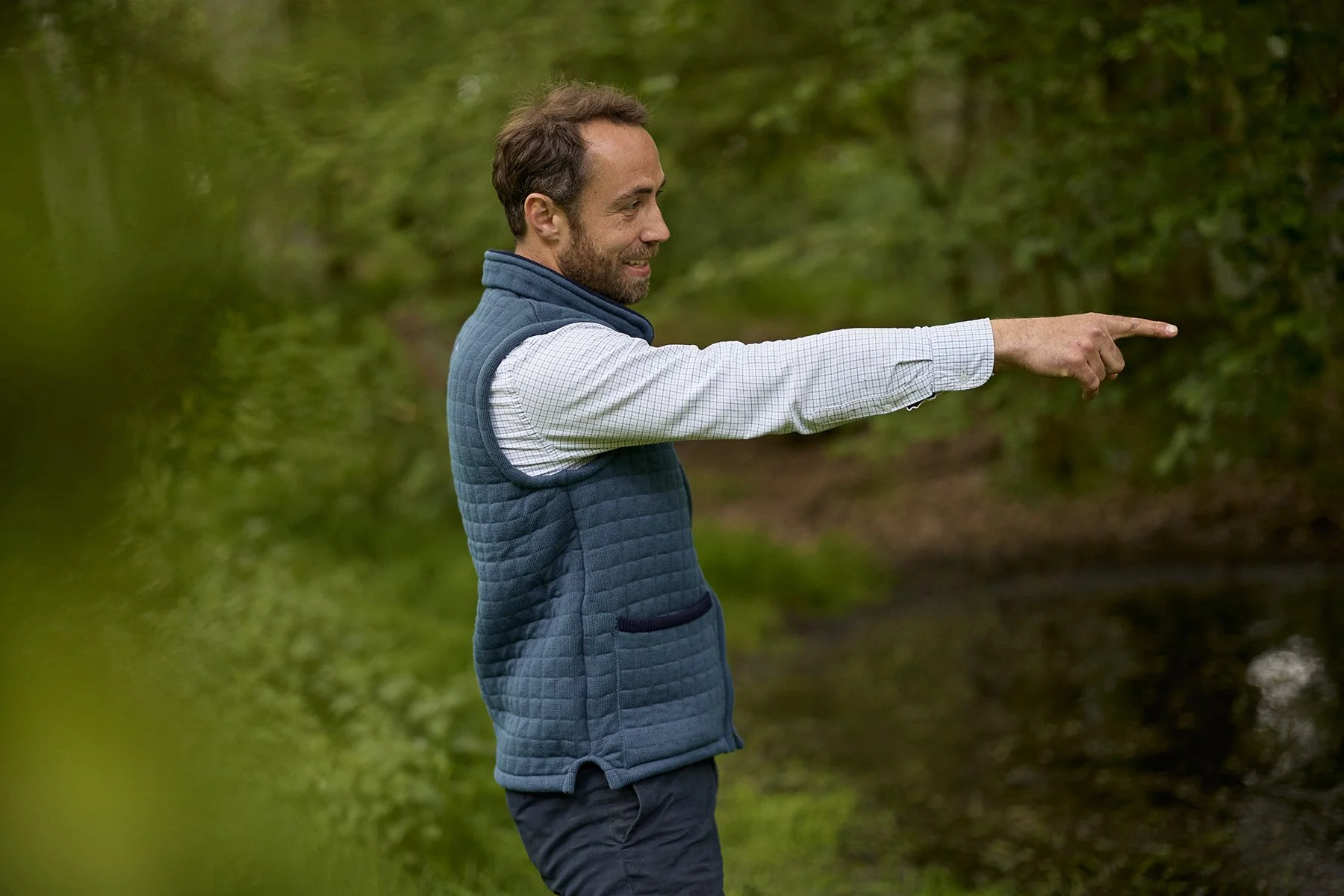 A man with brown hair and a beard, wearing a blue quilted vest and a white checkered shirt, points towards something in a wooded area next to a river.