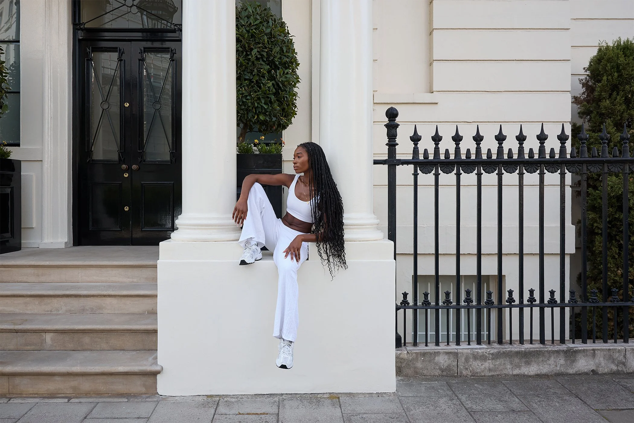 A woman with long curly hair sitting on a ledge outside a white building with black fence and stairs, wearing white sporty outfit.