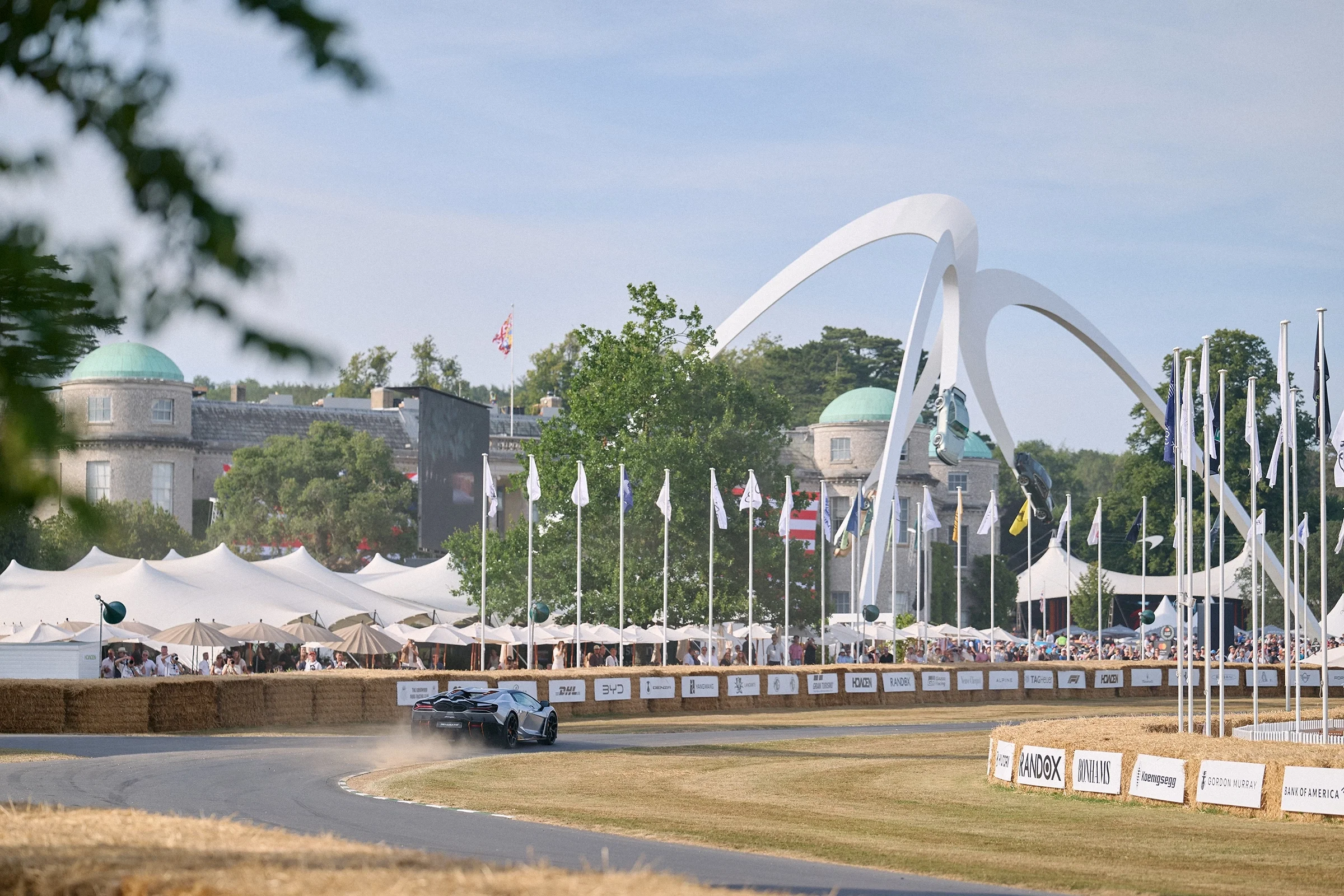 Supercar accelerating up the Goodwood Festival of Speed 2026 hill climb with Goodwood House and hospitality suites in the background.