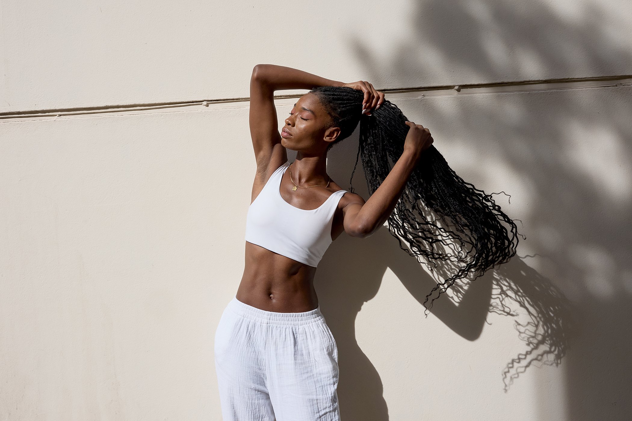 A woman with dark skin and curly hair standing against a beige wall, stretching her arms and eyes closed.