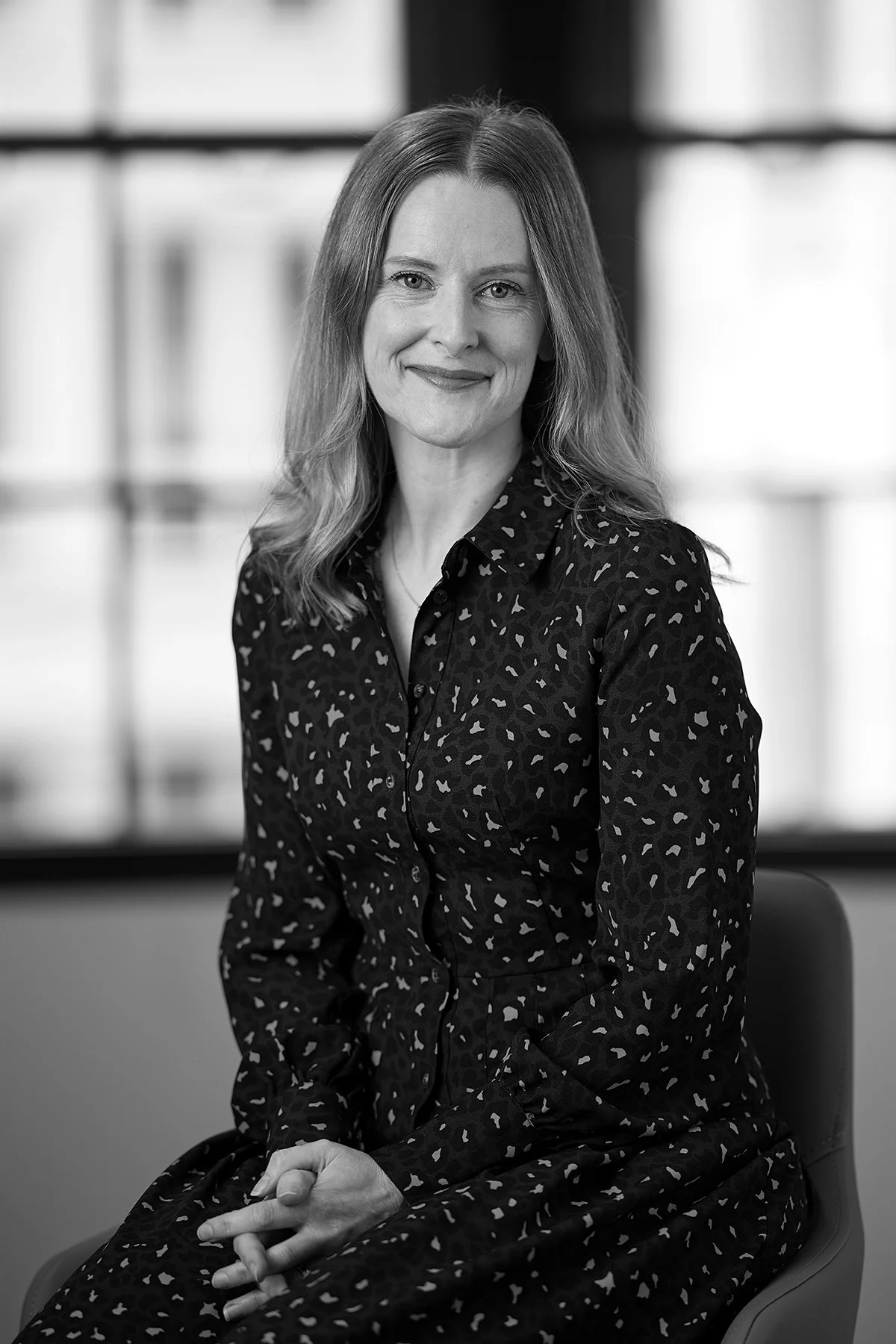 A woman with wavy hair, wearing a patterned dress, sitting and smiling in a professional setting with windows in the background.