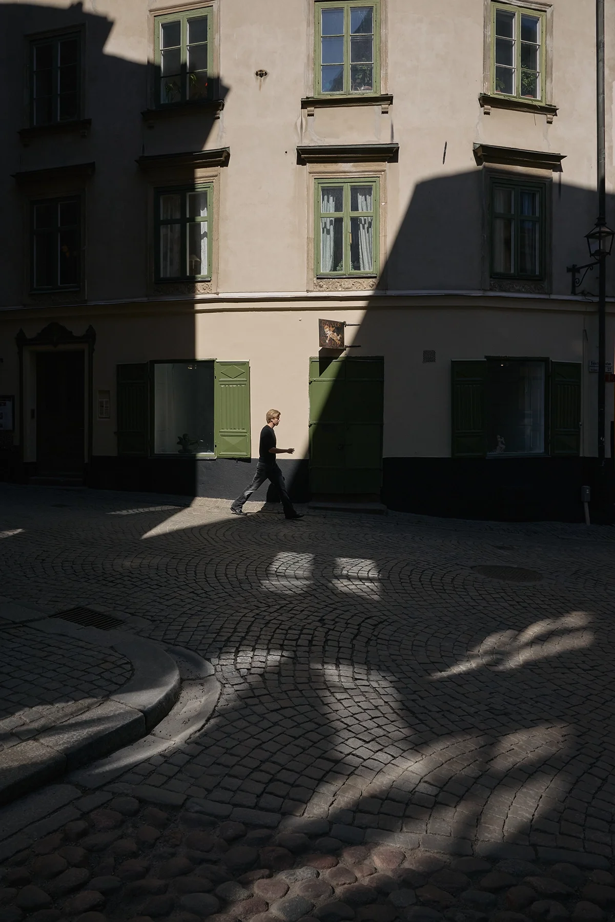 Intersection of narrow old streets with strong light and shadow contrast, featuring a single person walking through the scene in a European city.