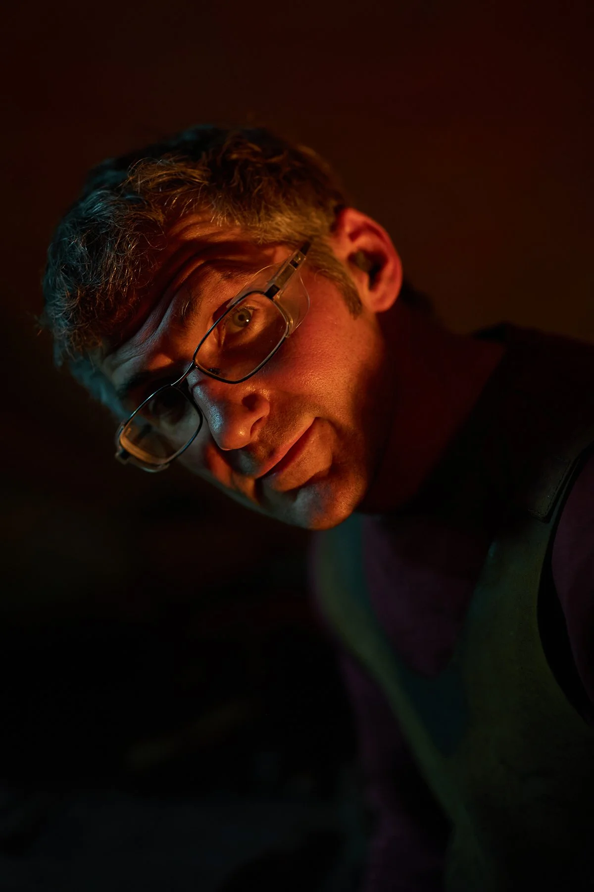 Close-up of a man with glasses looking intently, with colorful lighting and dark background.