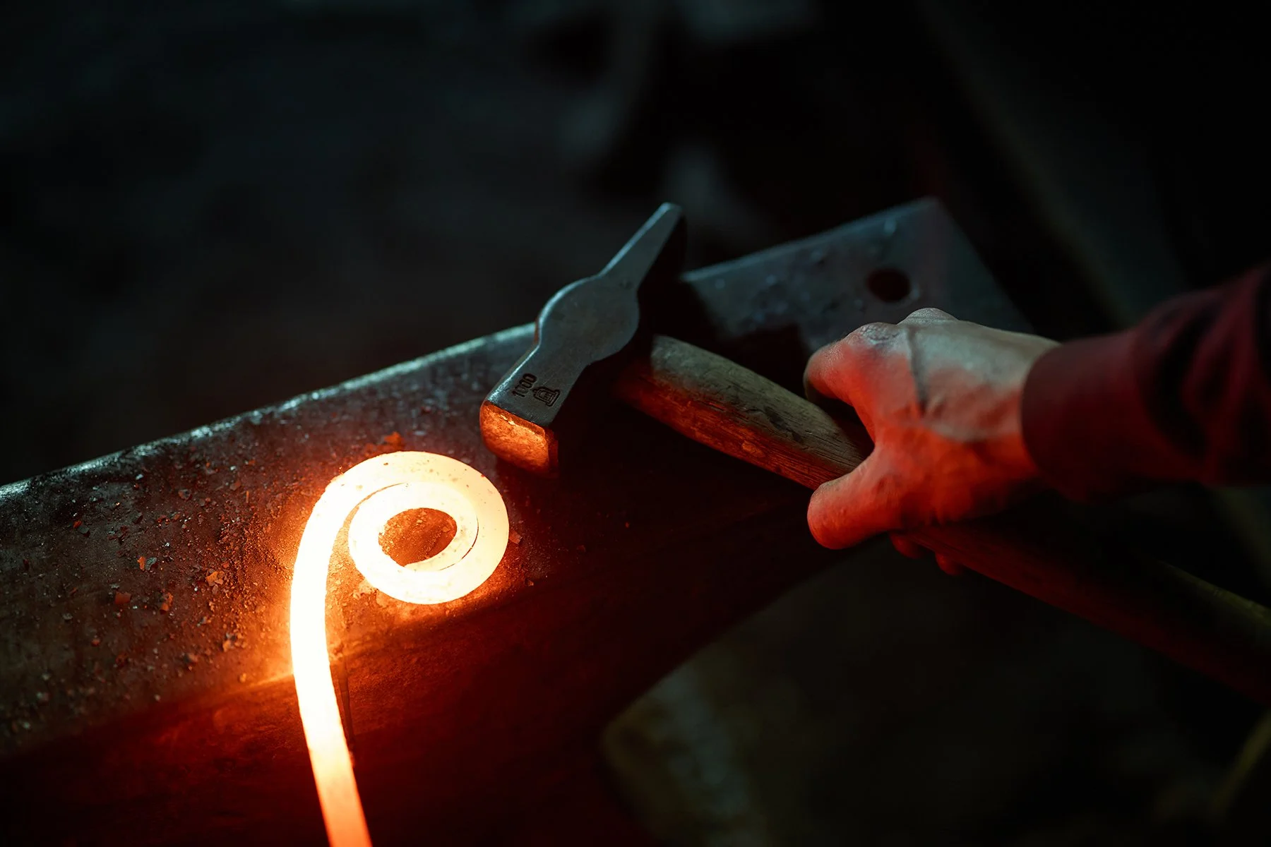 Close-up commercial photography of a blacksmith's hand with a hammer hitting glowing red metalwork, capturing the authentic forging process for branding assets.