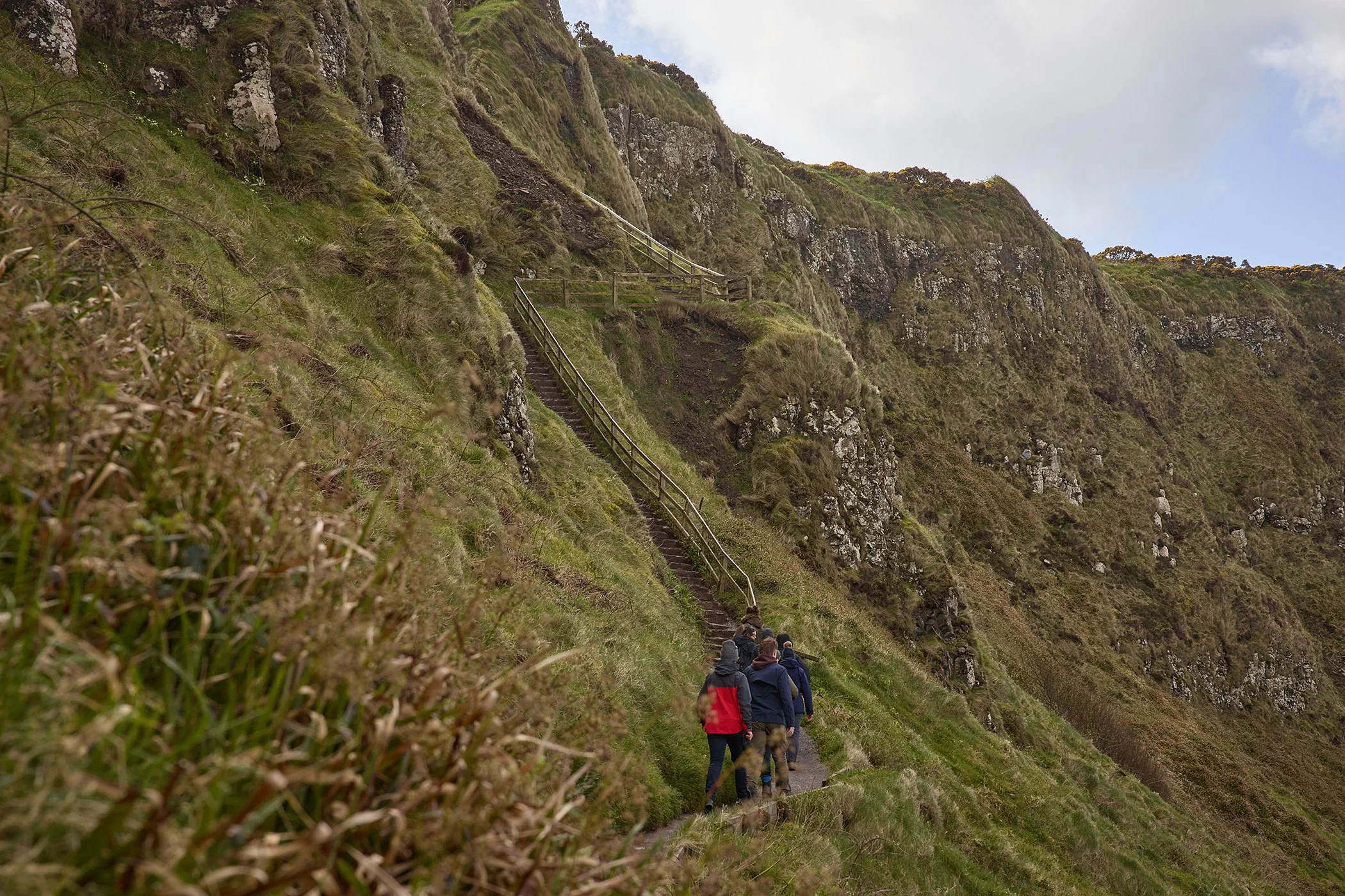 A group of hikers walking up a narrow, steep trail with stairs along a grassy hillside or cliff side.
