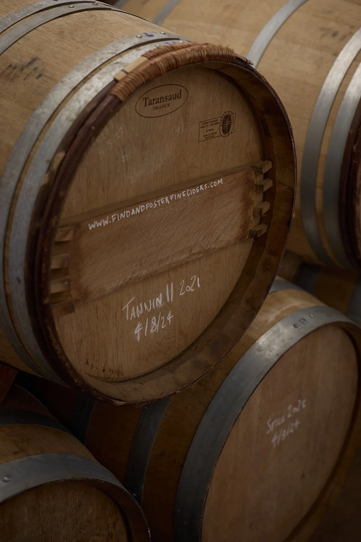 Wooden wine barrels stacked horizontally in a winery, with markings and website URL written on the back of one barrel.