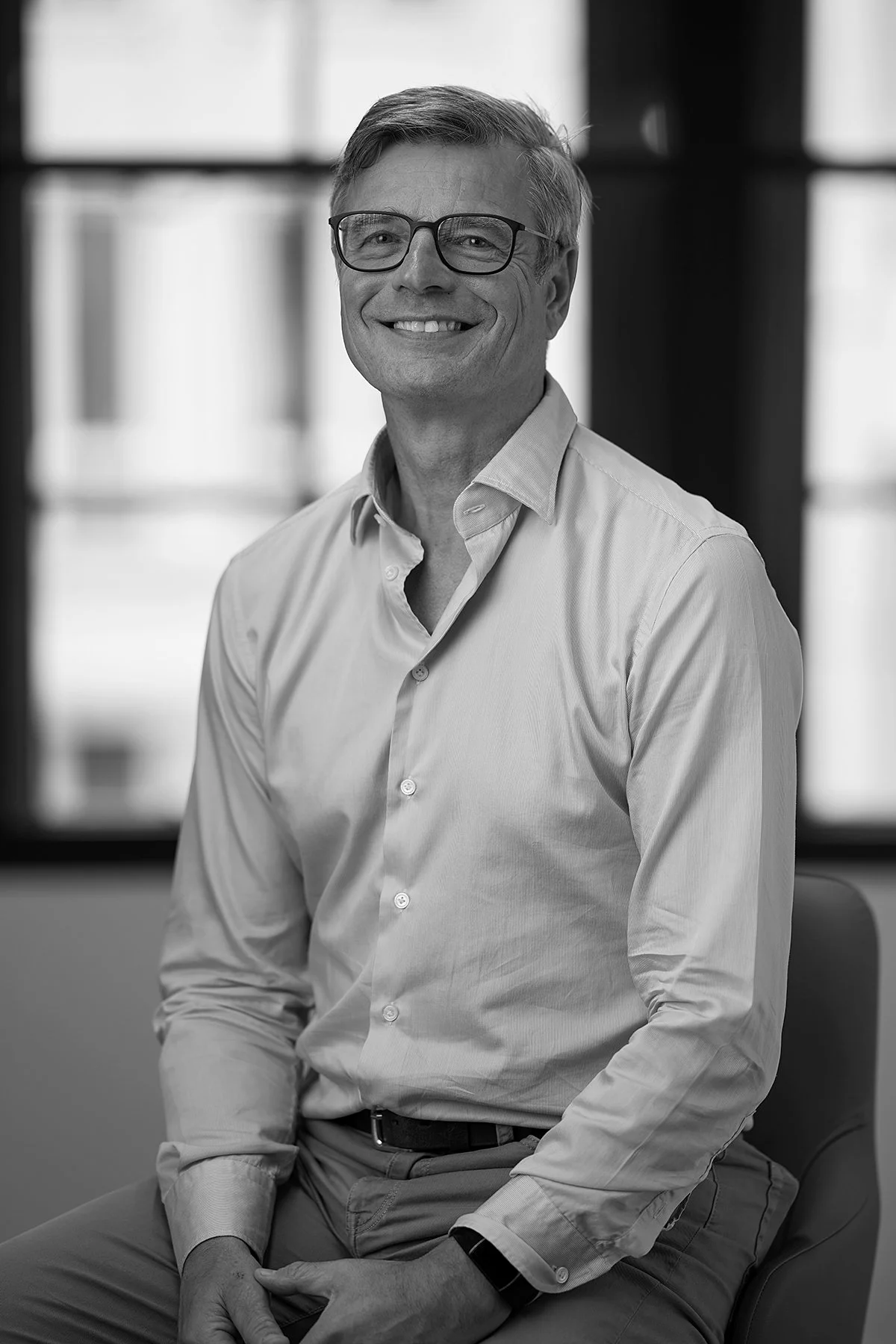 Black-and-white portrait of a smiling middle-aged man wearing glasses, a light-colored dress shirt, sitting in an office setting.
