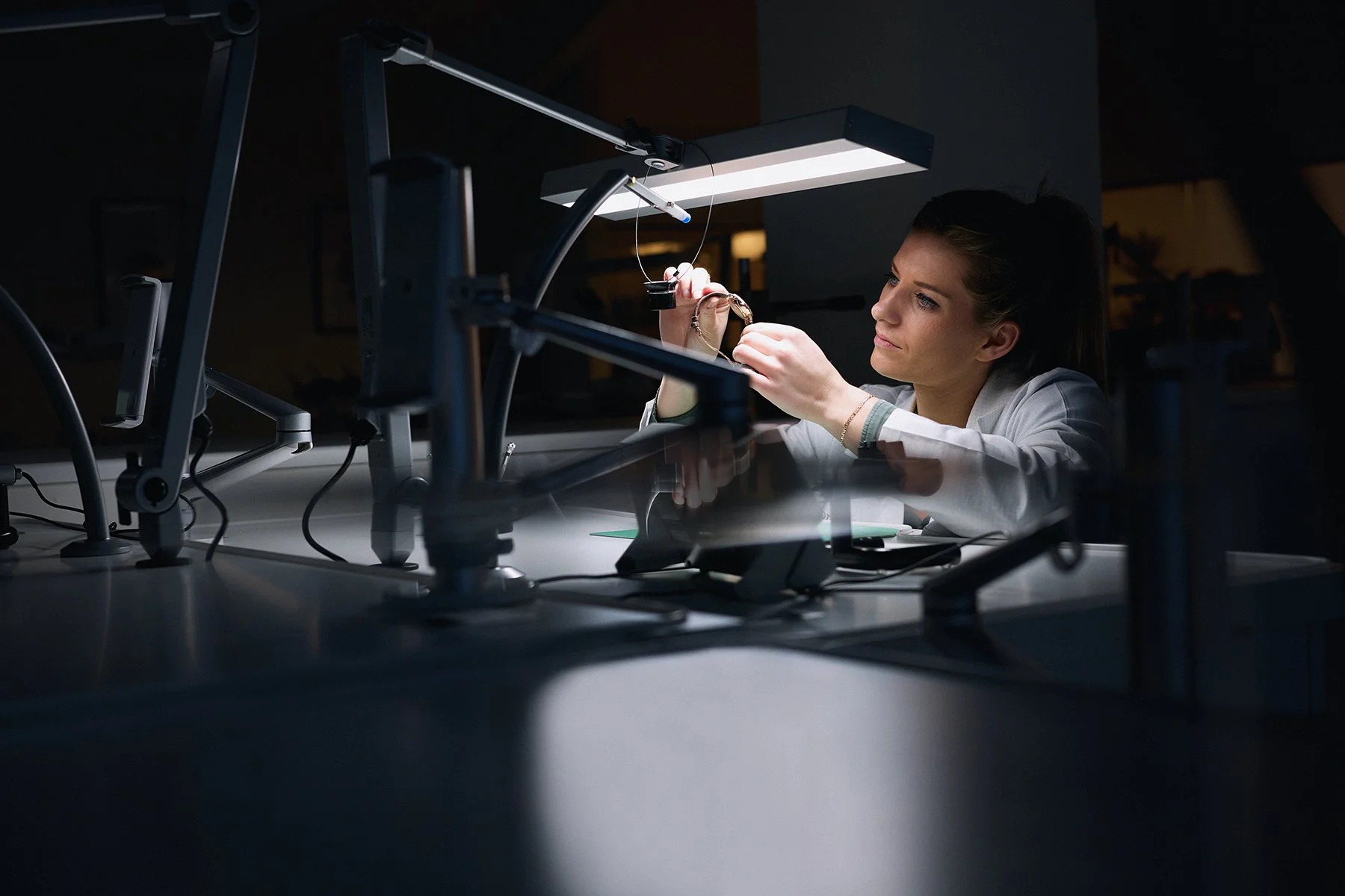 A woman working meticulously at a scientific laboratory bench, illuminated by a bright desk lamp, surrounded by equipment and tools.