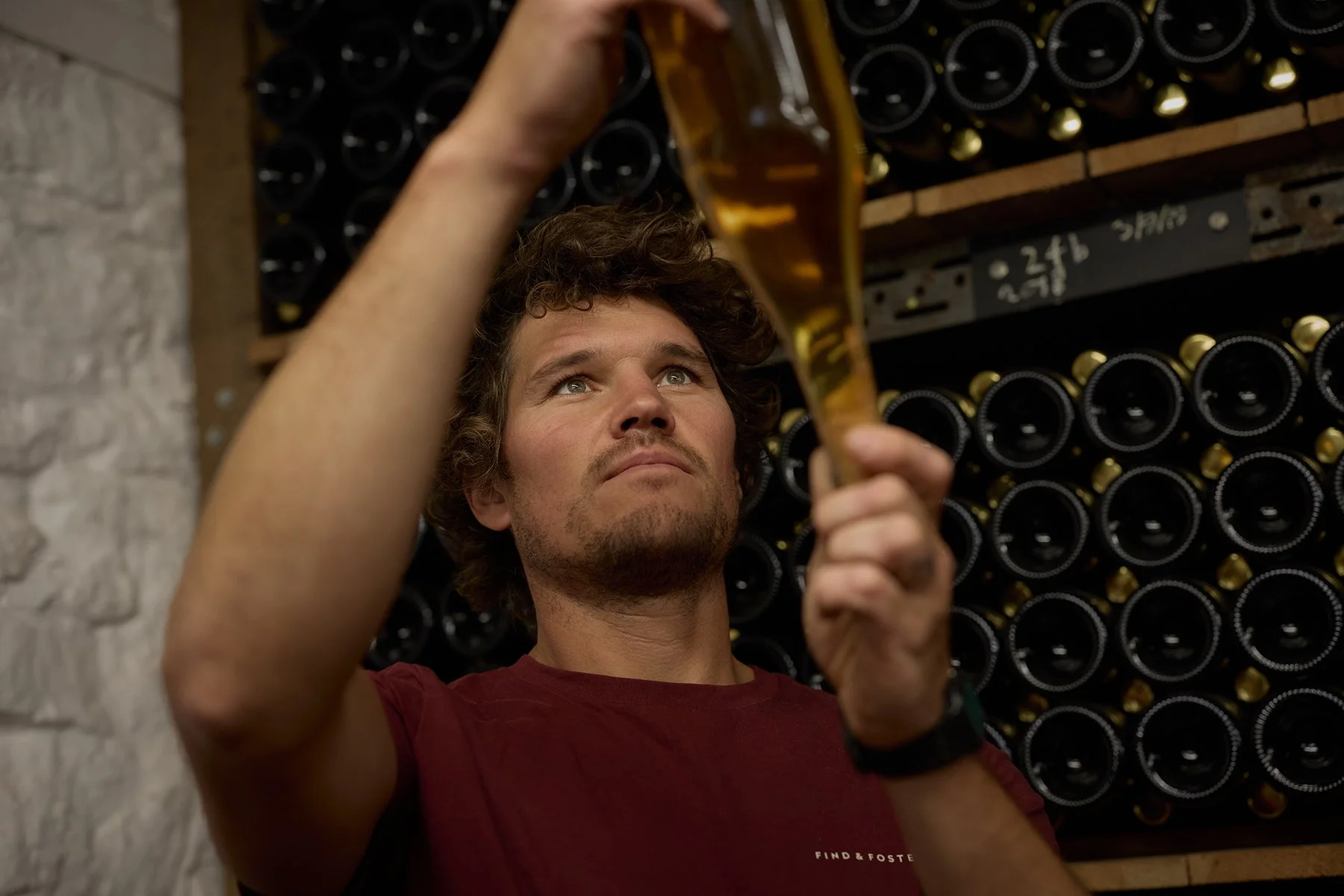 A young man with curly hair and a red shirt pouring wine from a bottle in a wine cellar.