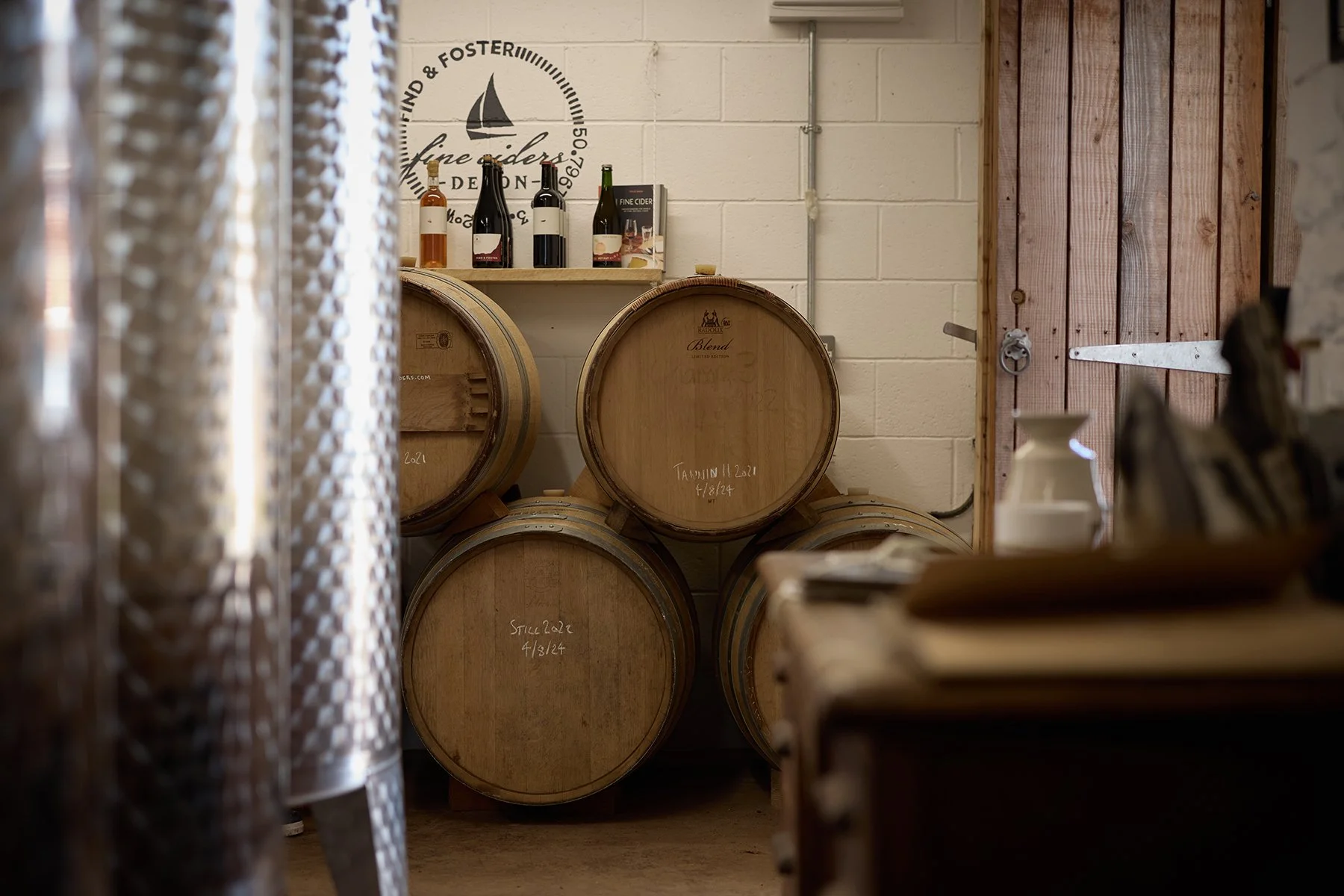 Row of wine barrels in a winery cellar with bottles and a shelf in the background.