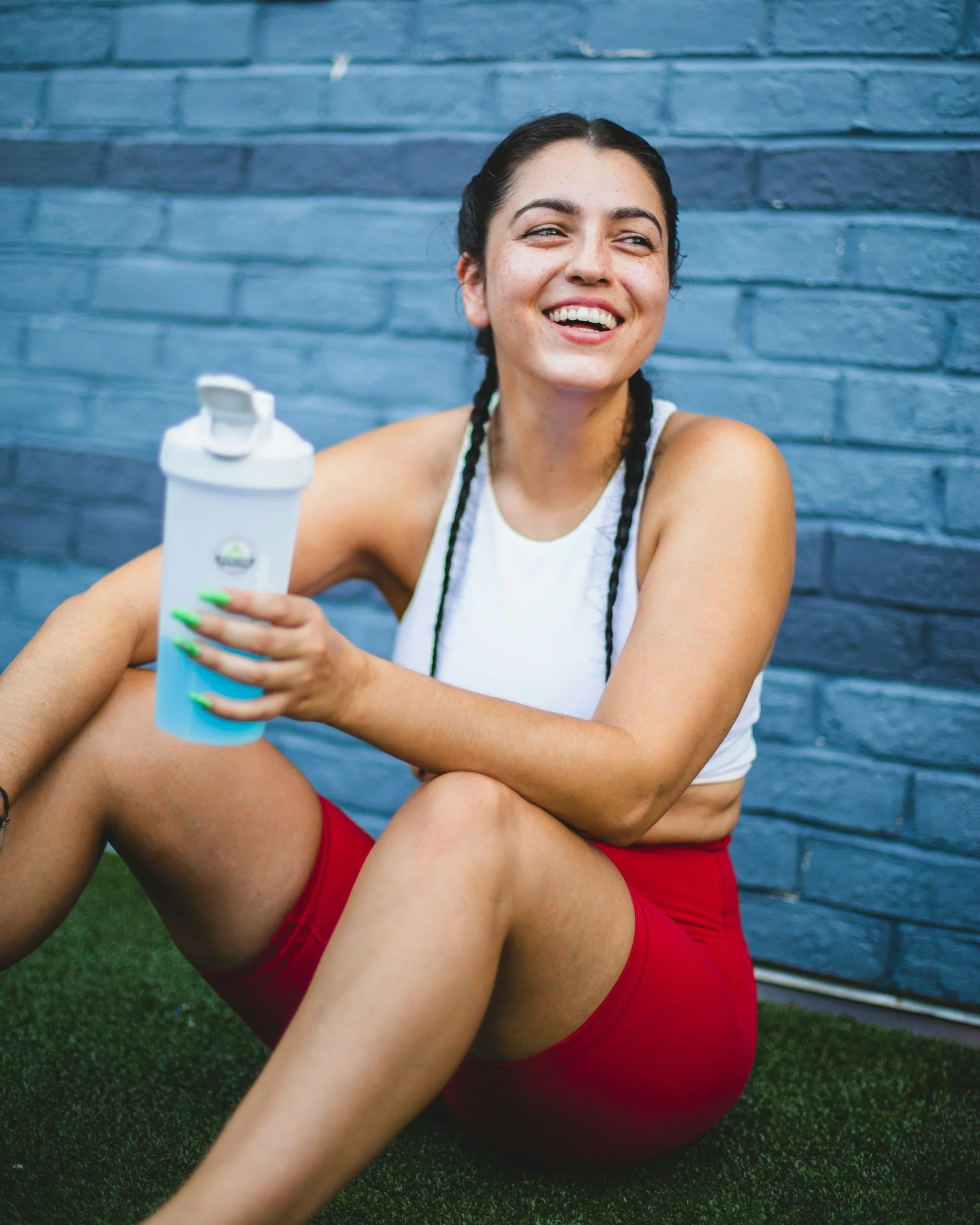 Young woman smiling, holding a water bottle, sitting on grass against a blue brick wall, wearing a white tank top and red shorts.
