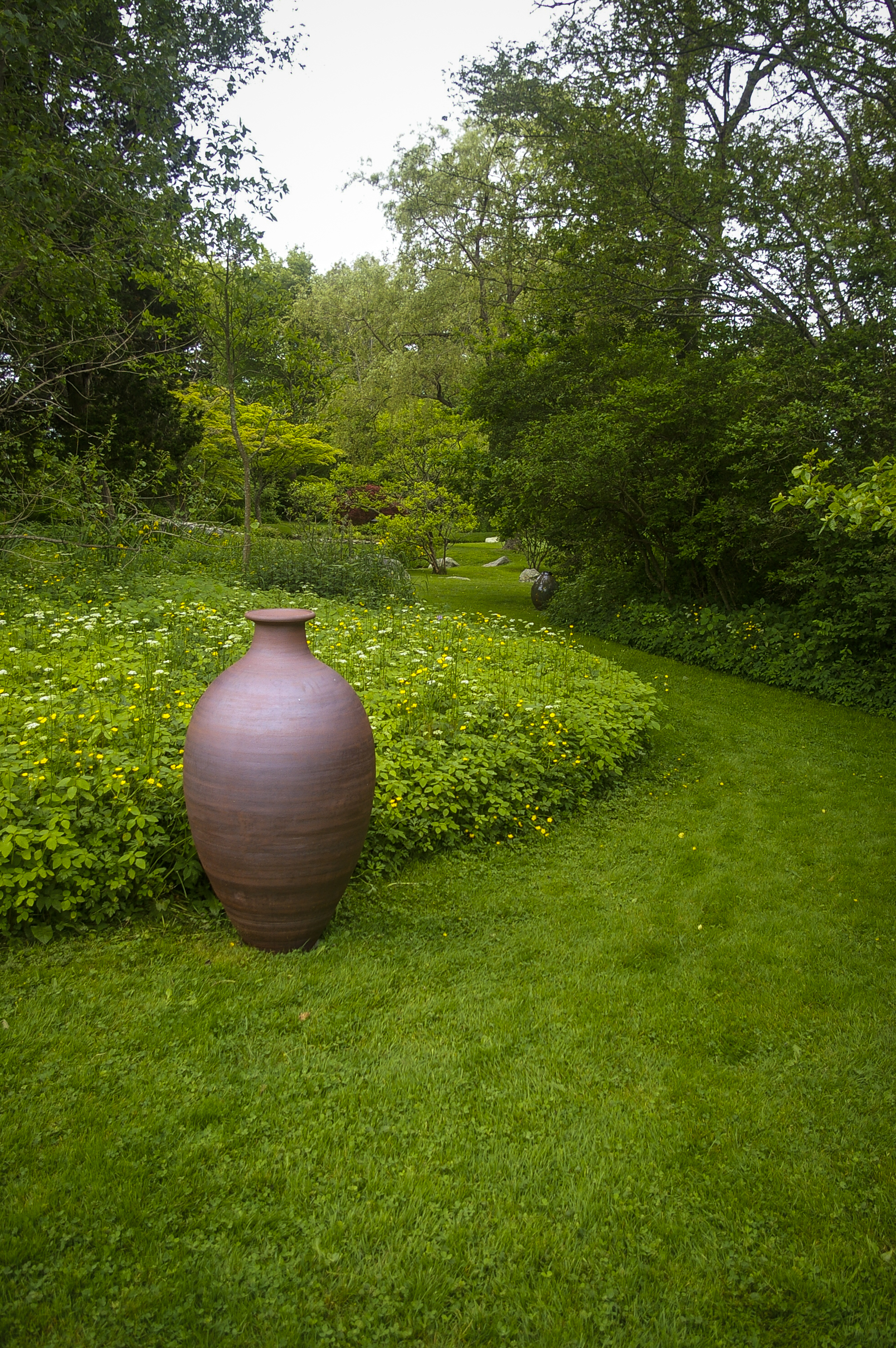 Tall ceramic urn displayed on grassy lawn surrounded by flowering shrubs