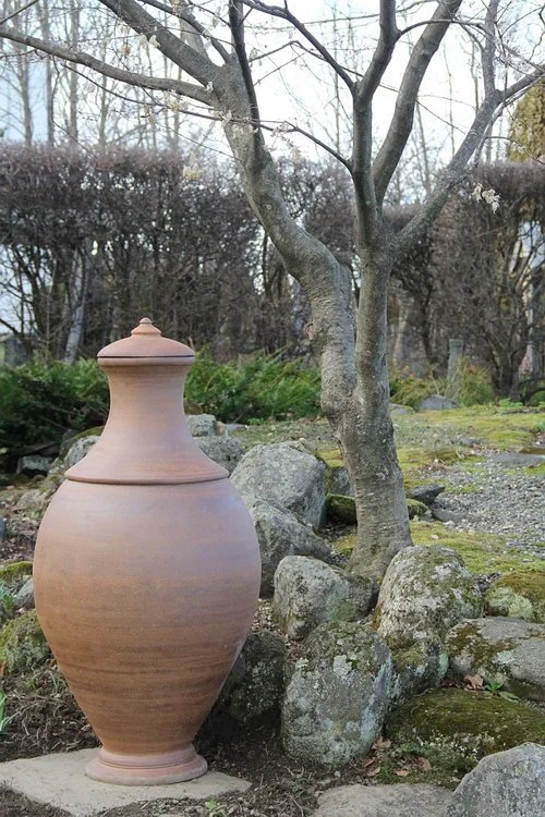 Decorative clay vessel positioned near bare trees and rocks in outdoor garden setting