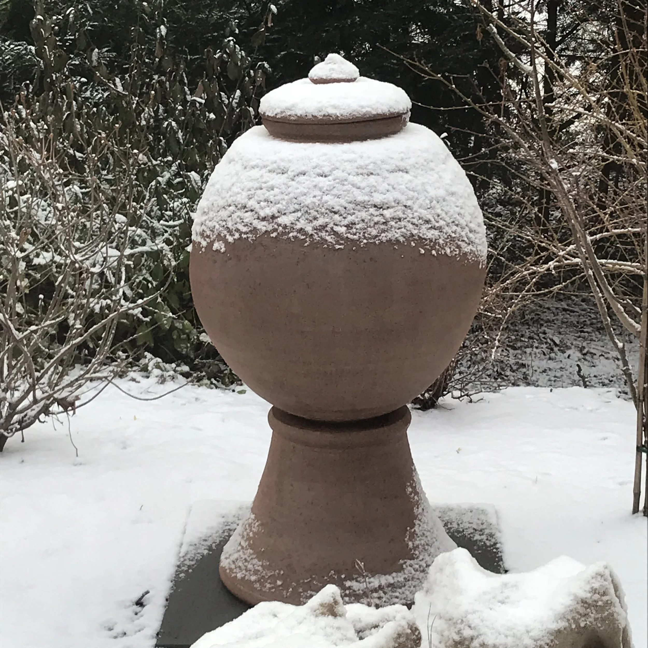 Large ceramic urn with lid on pedestal, partially covered in snow, in a winter garden setting.