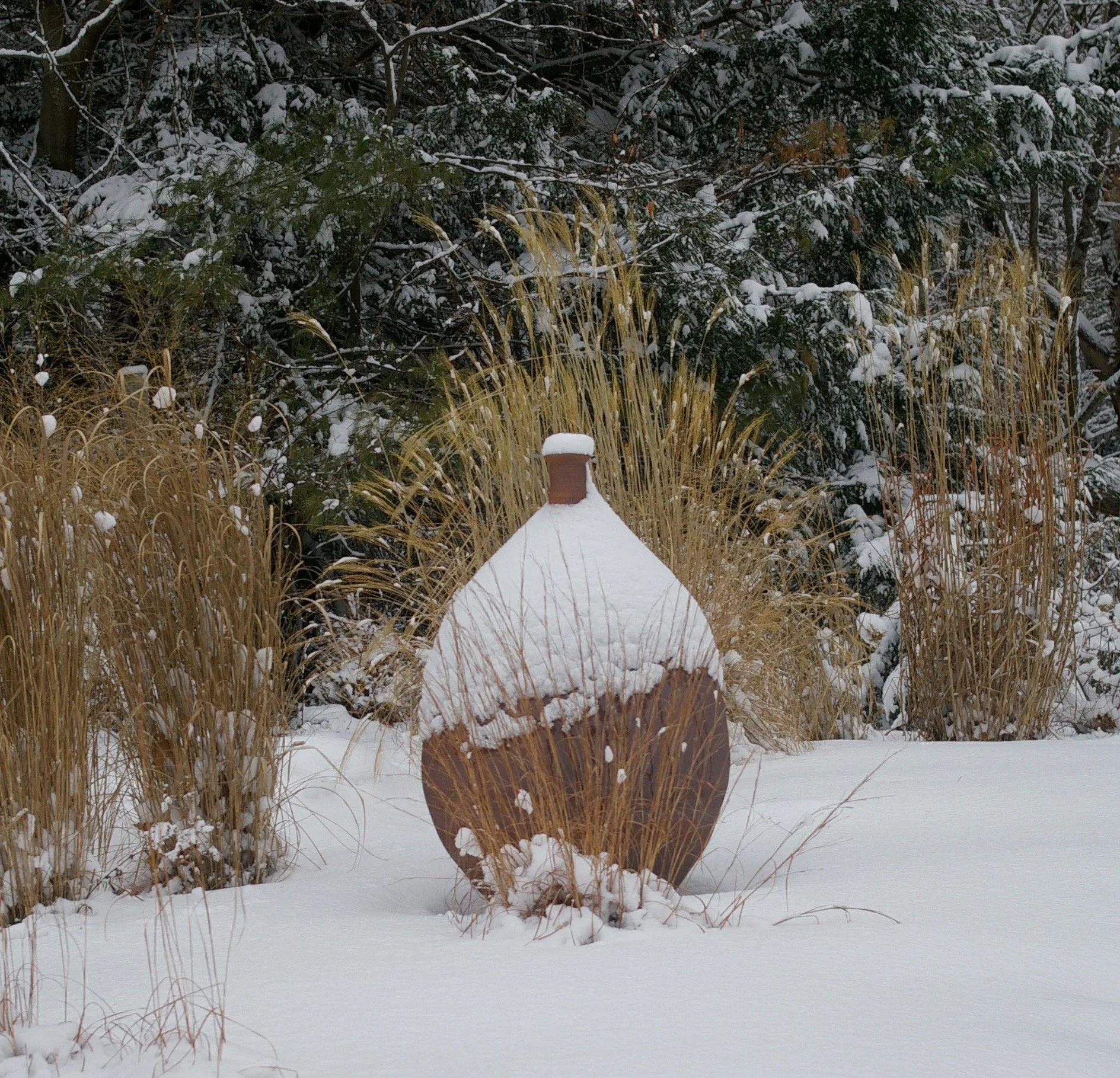 Round ceramic vase partially covered with snow, surrounded by ornamental grasses and evergreen trees in winter garden.