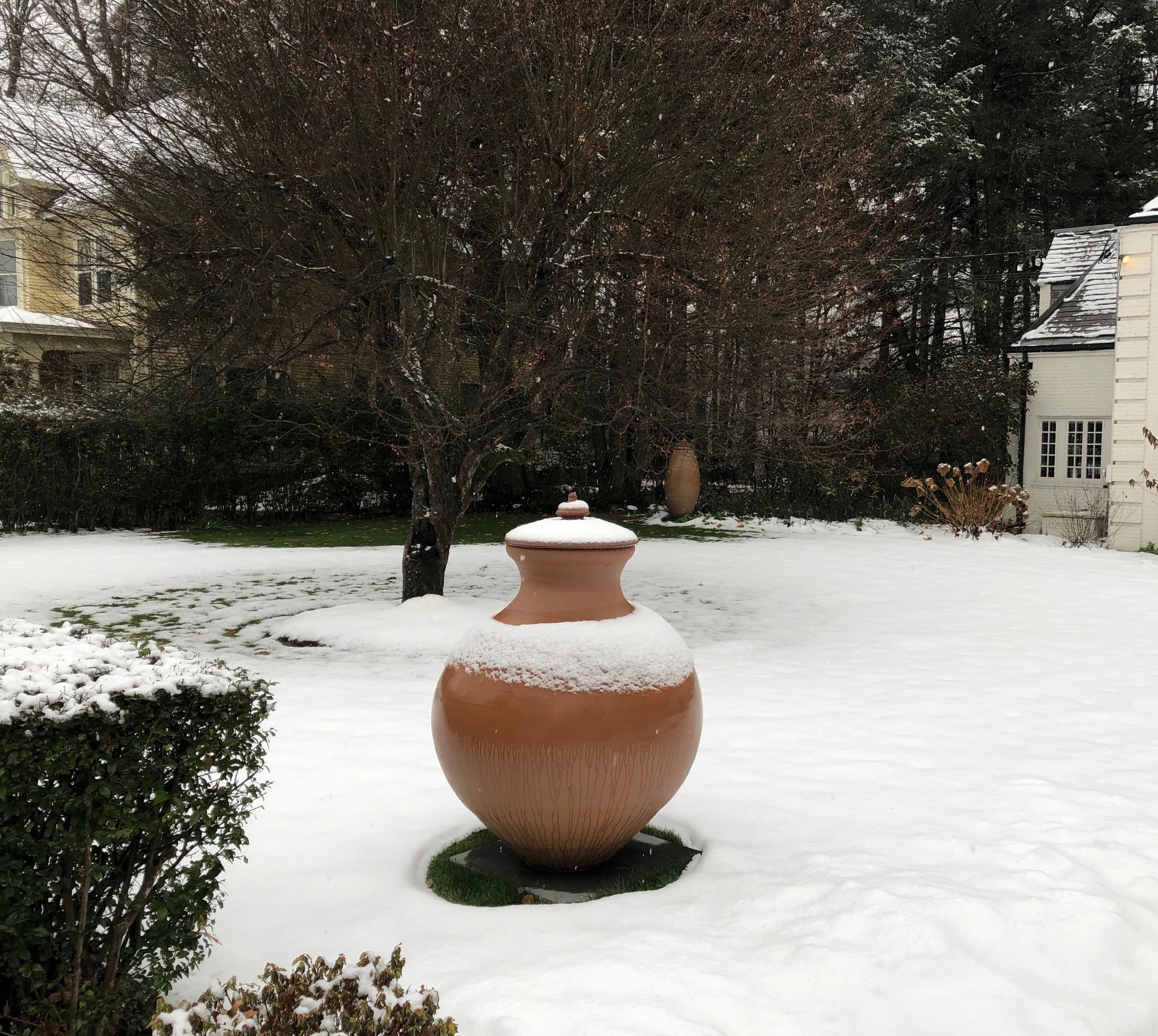 Snow-covered yard with large ceramic vase in foreground, second vase near tree, winter garden scene with houses in background.