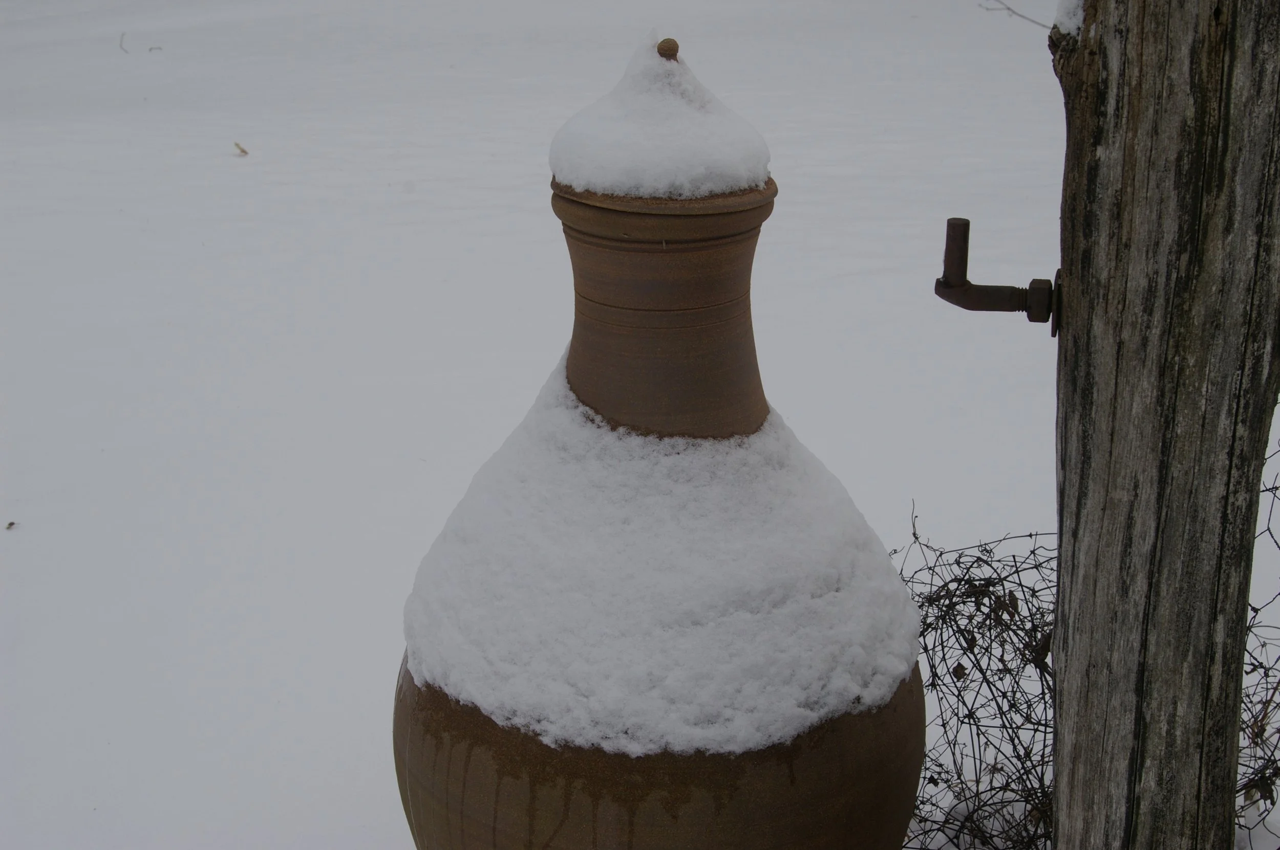 Large clay pot topped with snow beside wooden post and spigot in a winter garden setting