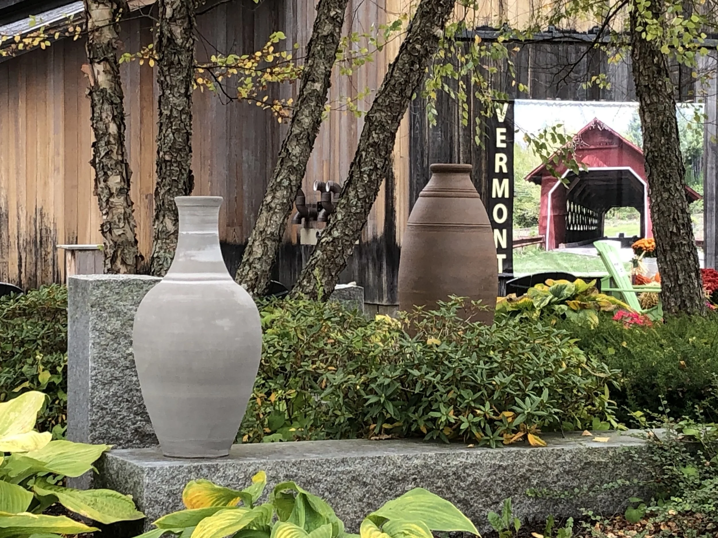 Outdoor display with two large pottery vases among green plants, rustic wooden building, and Vermont sign with covered bridge view.