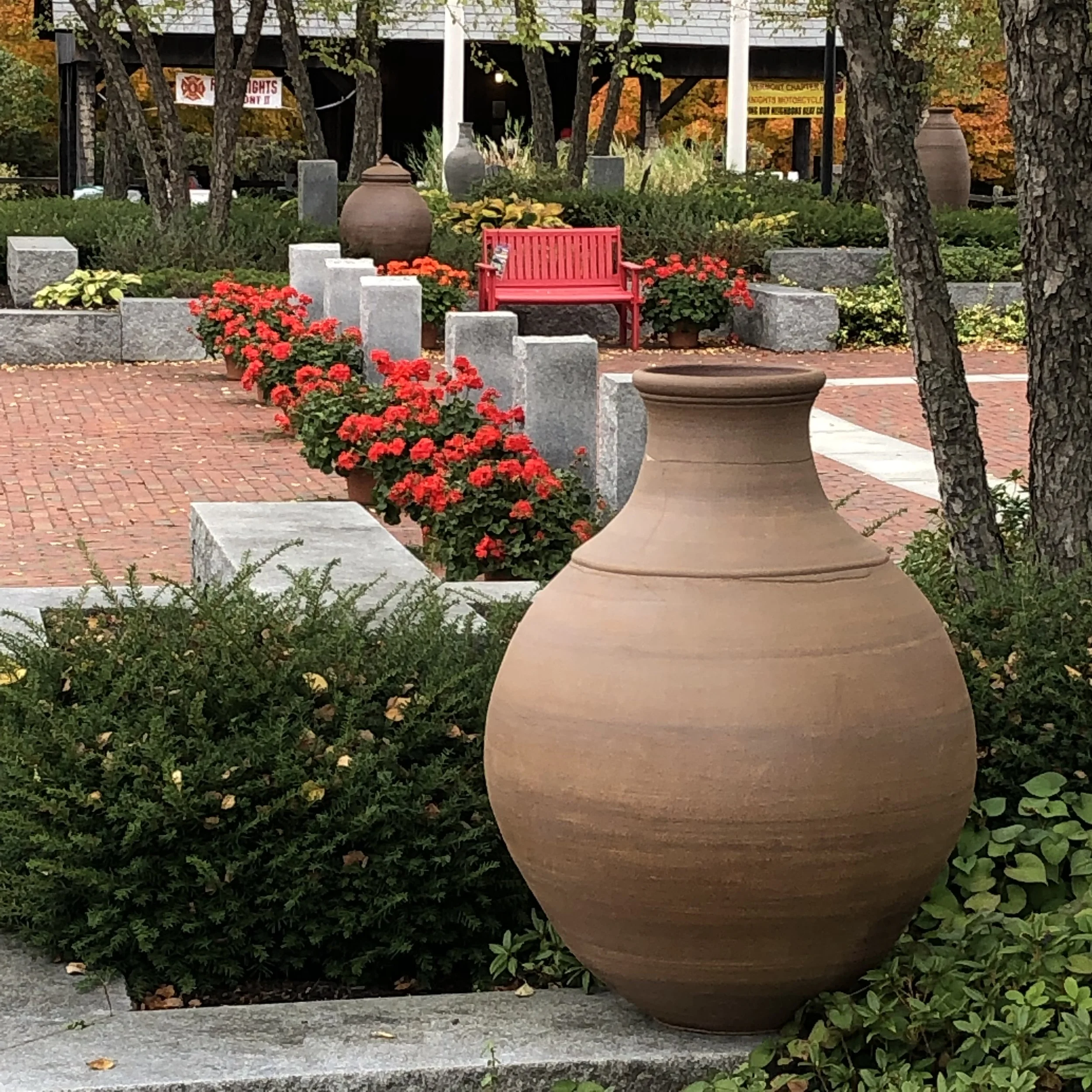 Large vessel in landscaped garden with red geraniums, stone pathway, bright red bench, and autumn foliage.