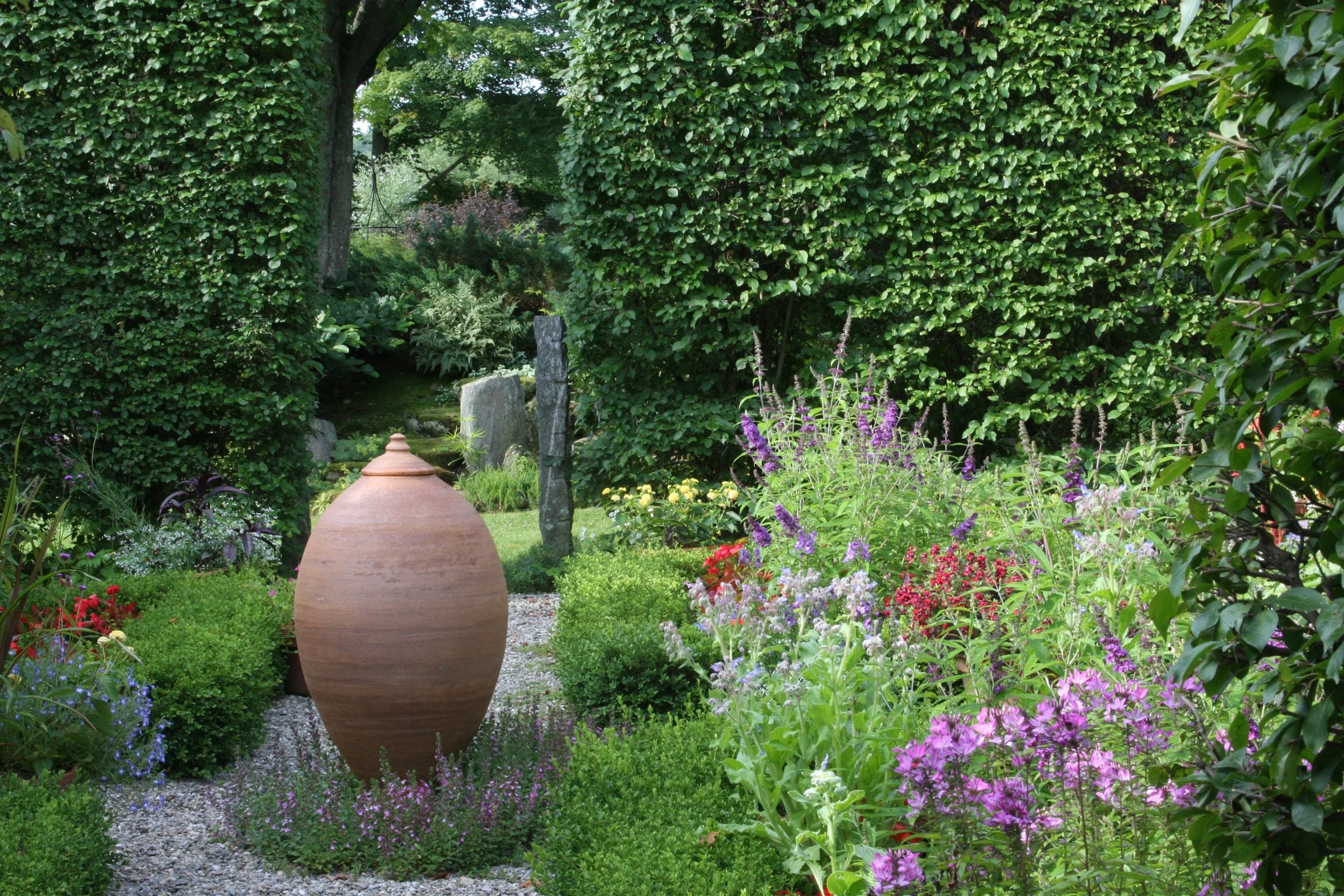 Mandorla sculpture in herb garden surrounded by vibrant flowers, terracotta urn, and lush greenery