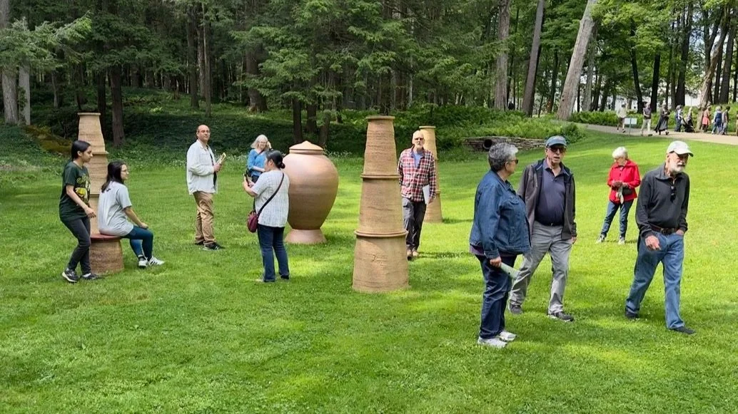 People viewing tall clay sculpture in outdoor art installation surrounded by greenery
