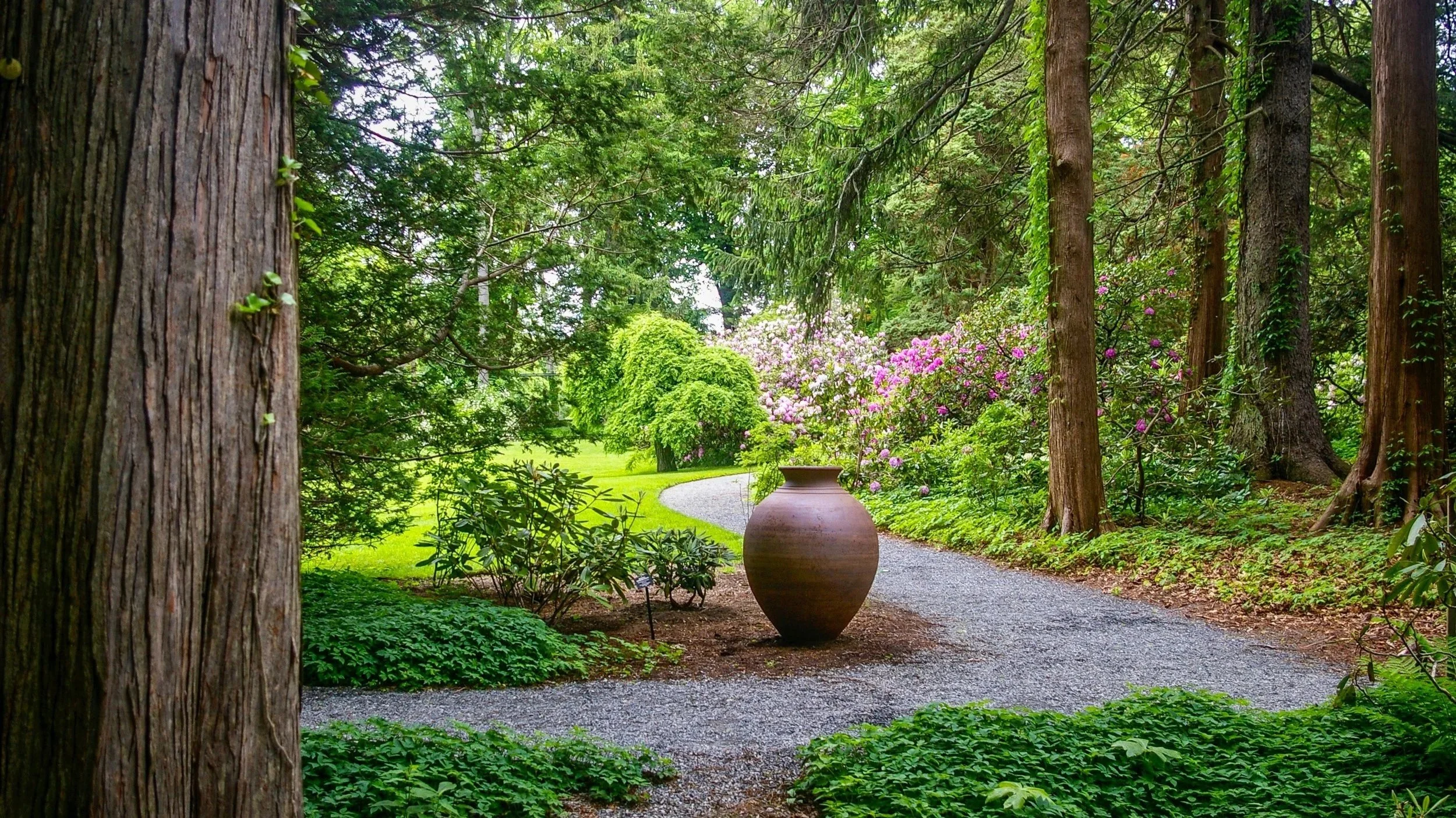 Handcrafted terracotta urn positioned along curved gravel path surrounded by lush greenery, trees, and flowering shrubs