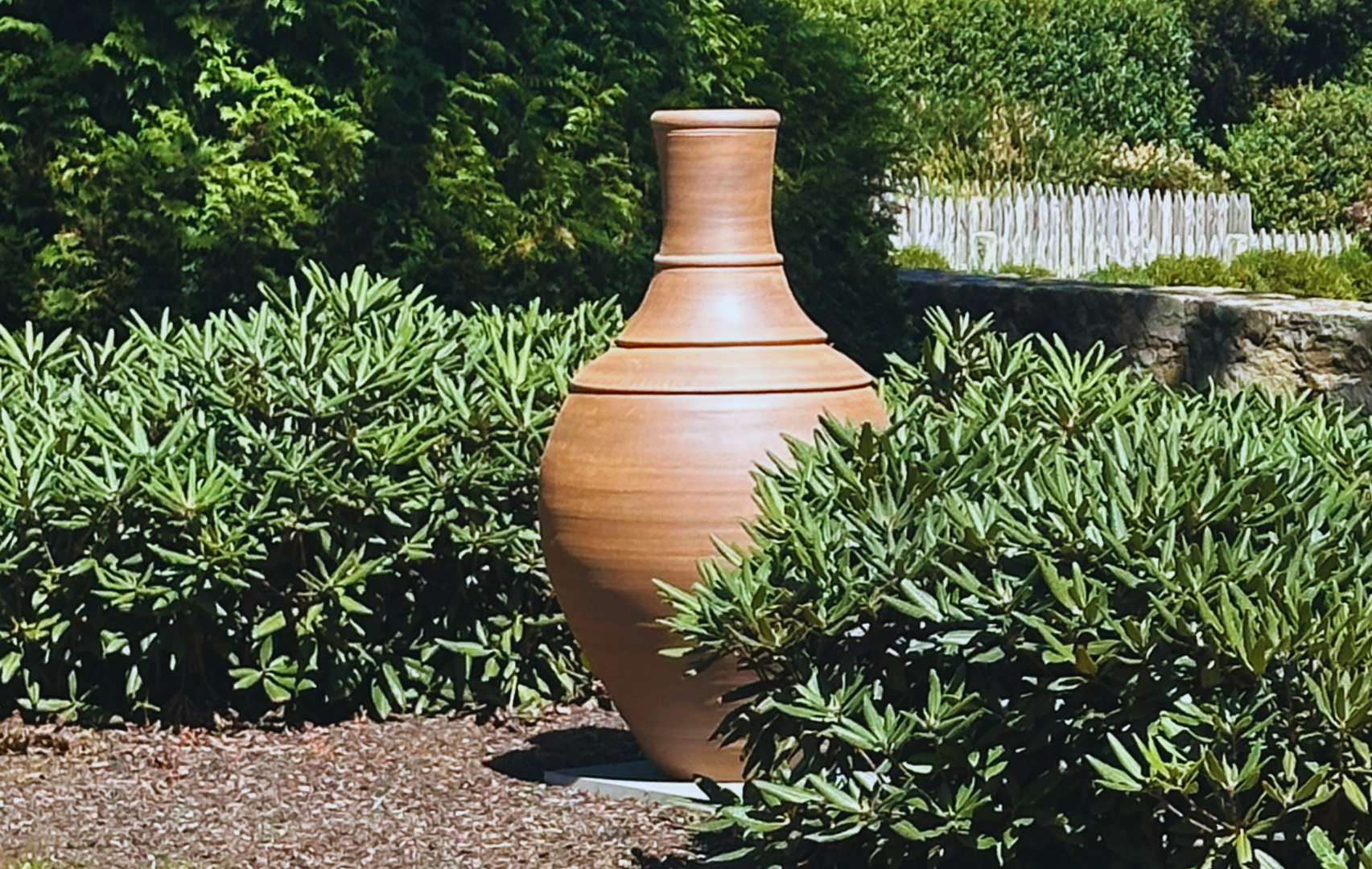  Handcrafted ceramic vase placed in lush green garden with stone wall, white picket fence, and surrounding trees