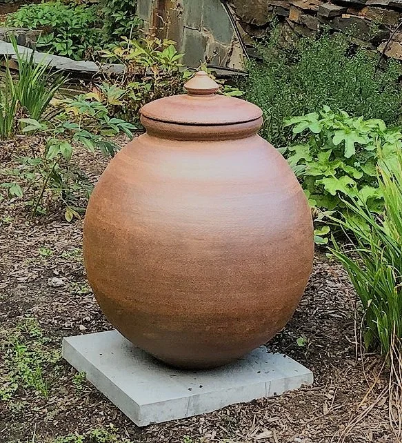 Terracotta garden vessel with lid displayed on concrete base surrounded by greenery