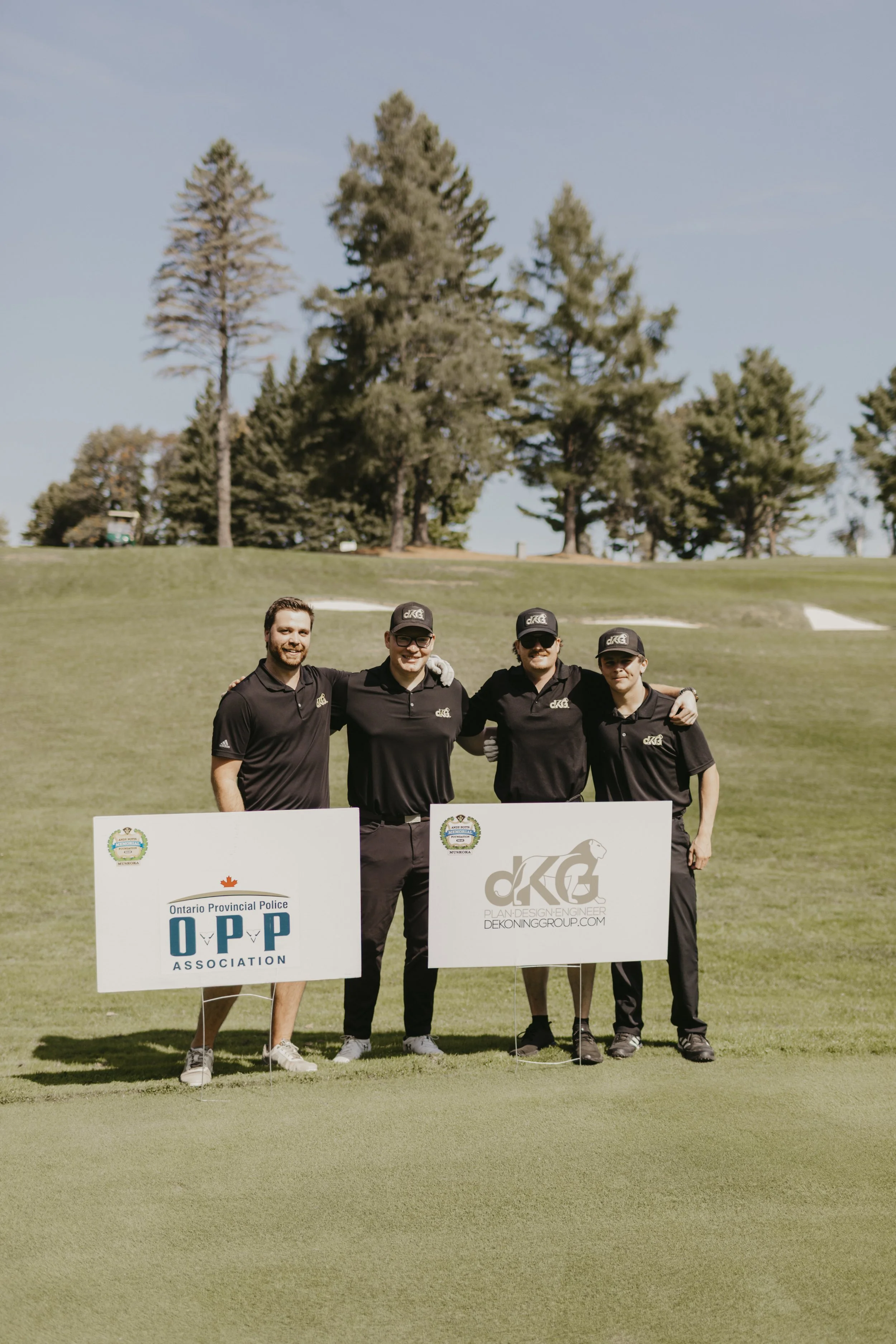 Four men standing on a golf course holding signs for the Ontario Provincial Police Association and a design group, with trees in the background.