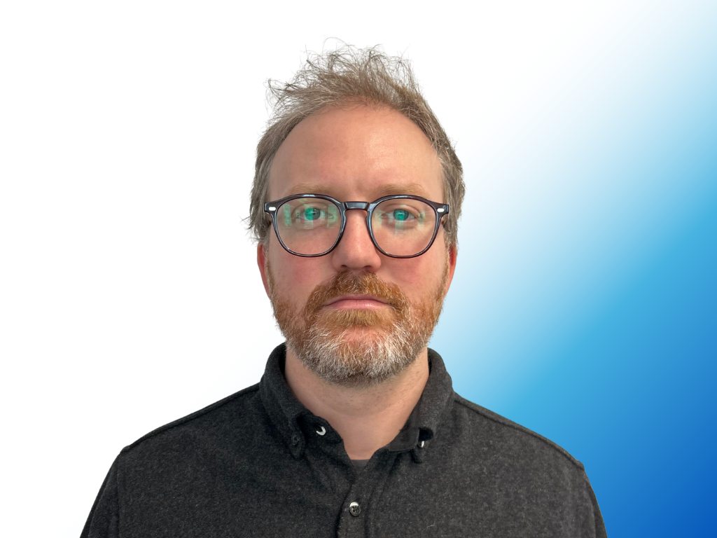 A man with glasses and a beard, wearing a black collared shirt, standing against a background that transitions from white to blue.