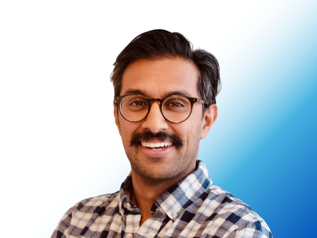 A smiling man with glasses, a mustache, and dark hair, wearing a plaid shirt.