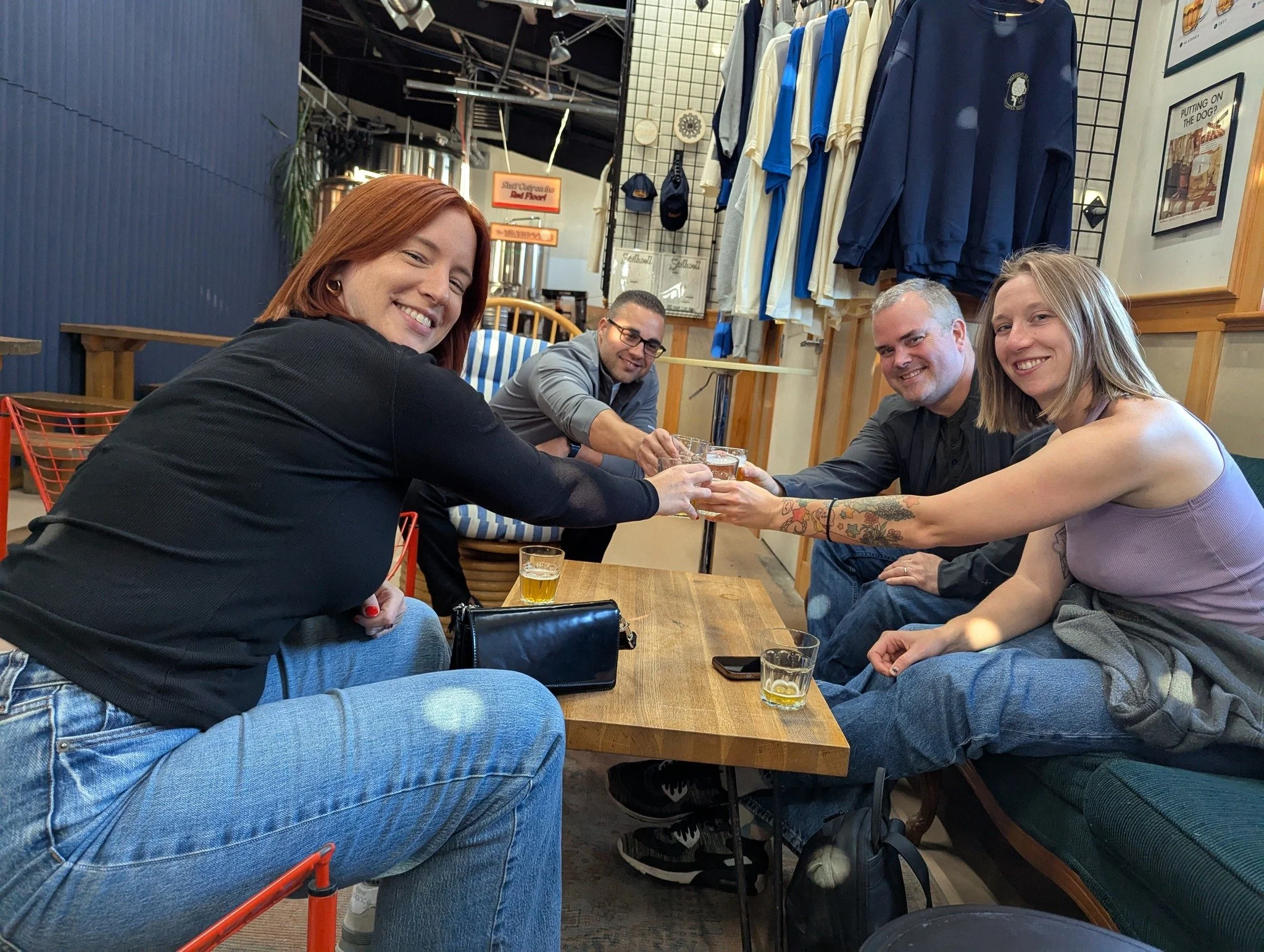 Four people sitting at a wooden table in a restaurant, smiling and toasting glasses of beer. The background shows clothing and framed pictures on the wall.