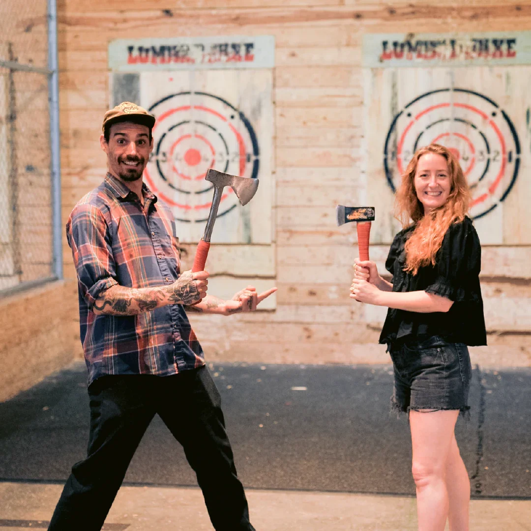 A man and woman holding axes at axe throwing range, smiling, with target boards in background.