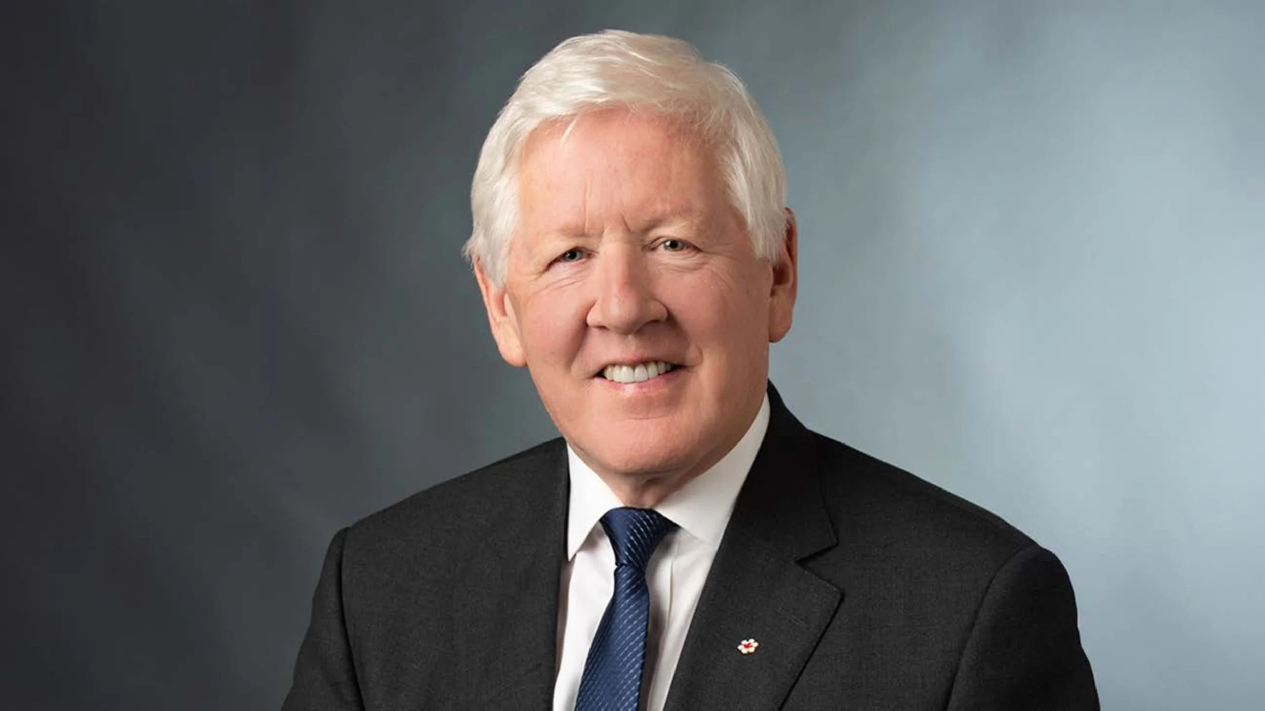 A professional headshot of a man, Bob Rae, with white hair, wearing a black suit, white shirt, and blue tie, smiling against a gradient background.