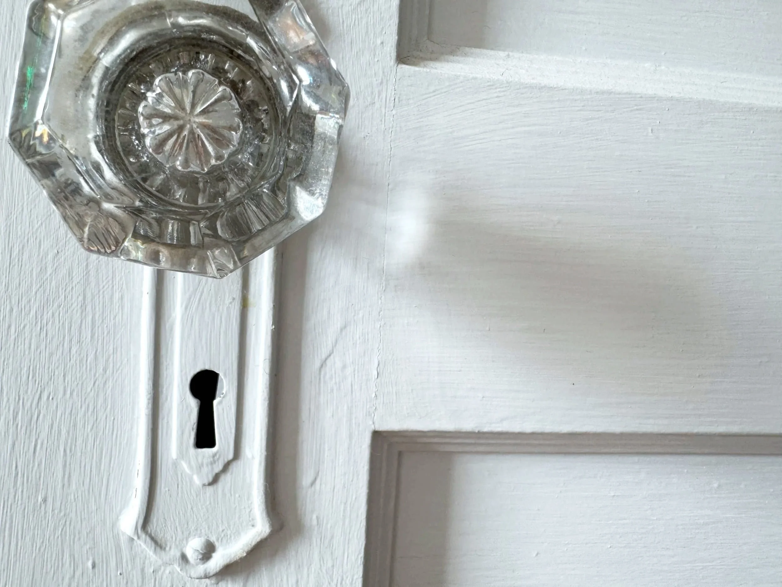Close-up of a vintage clear glass doorknob with a decorative pattern, mounted on a white wooden door with a keyhole below it.