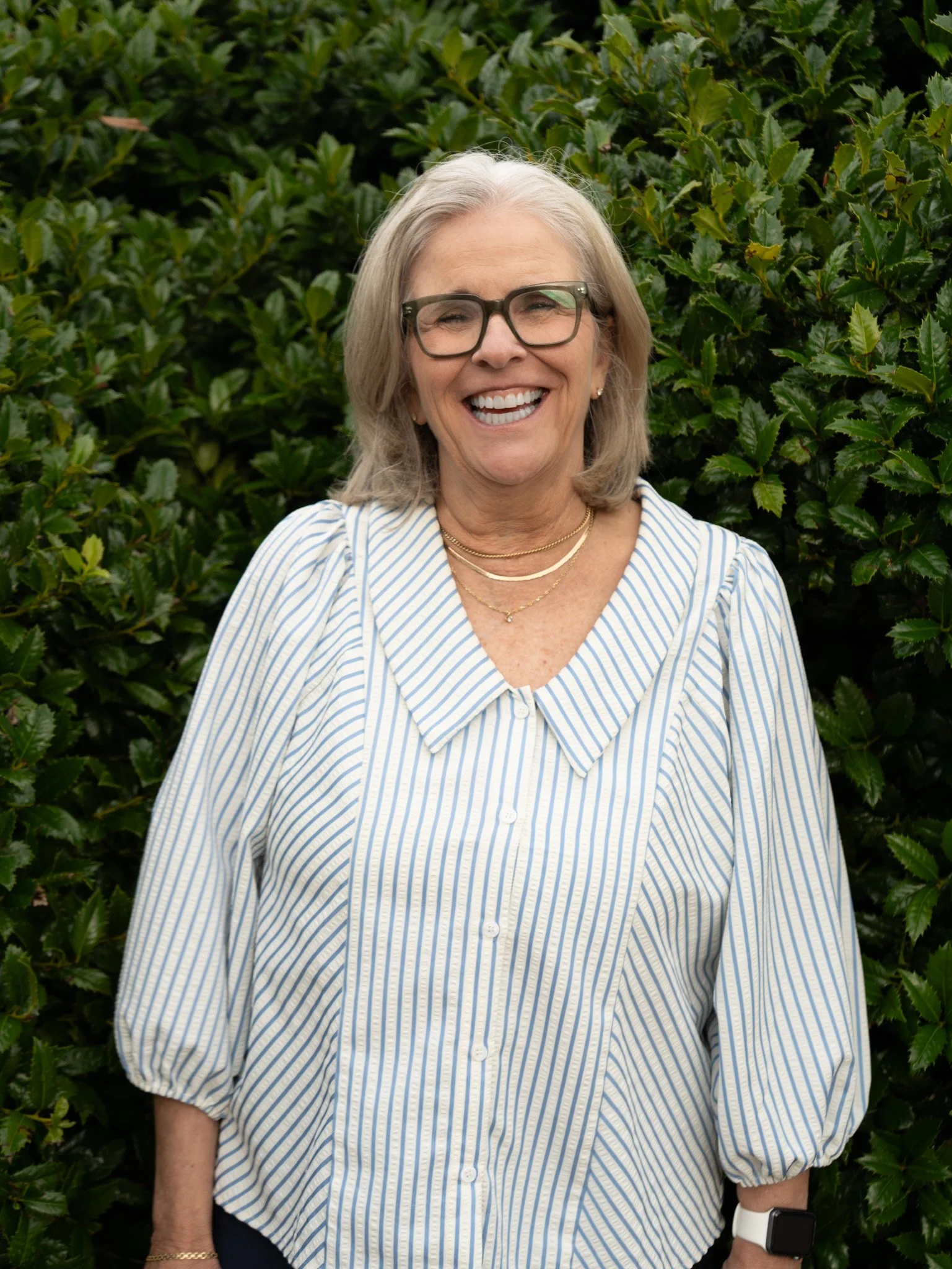 A woman with short gray hair, glasses, and earrings smiling in front of a decorative wrought iron and wooden wall.