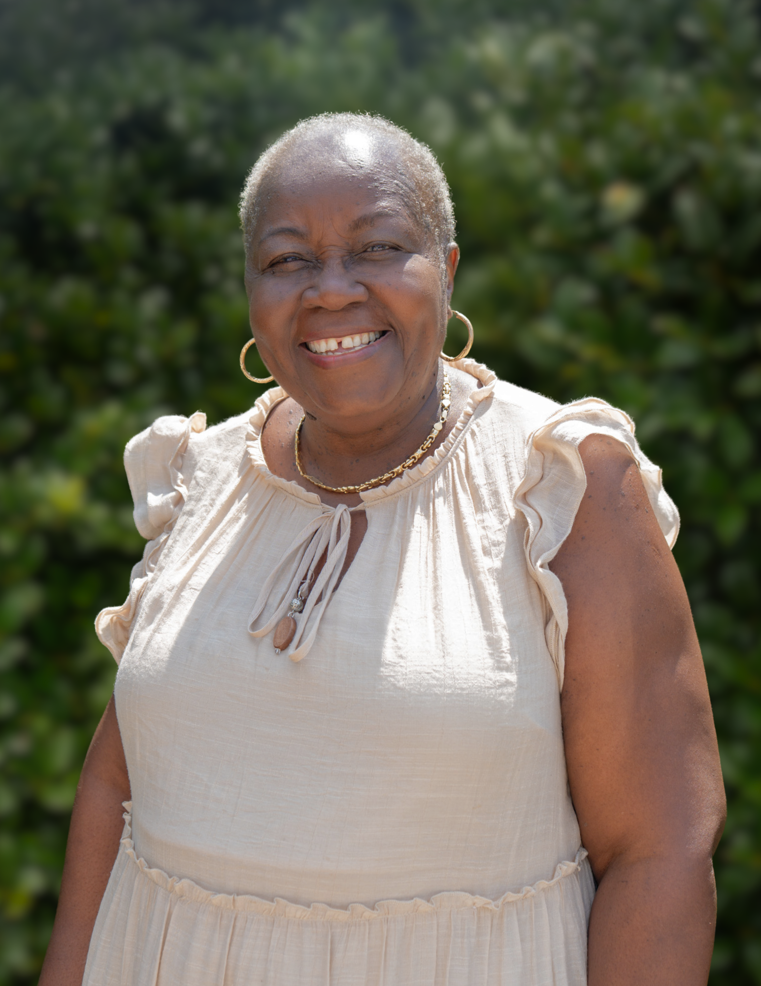 A smiling elderly woman with short gray hair wearing a multicolored sweater and hoop earrings, sitting in a room with yellow walls and wooden decor.
