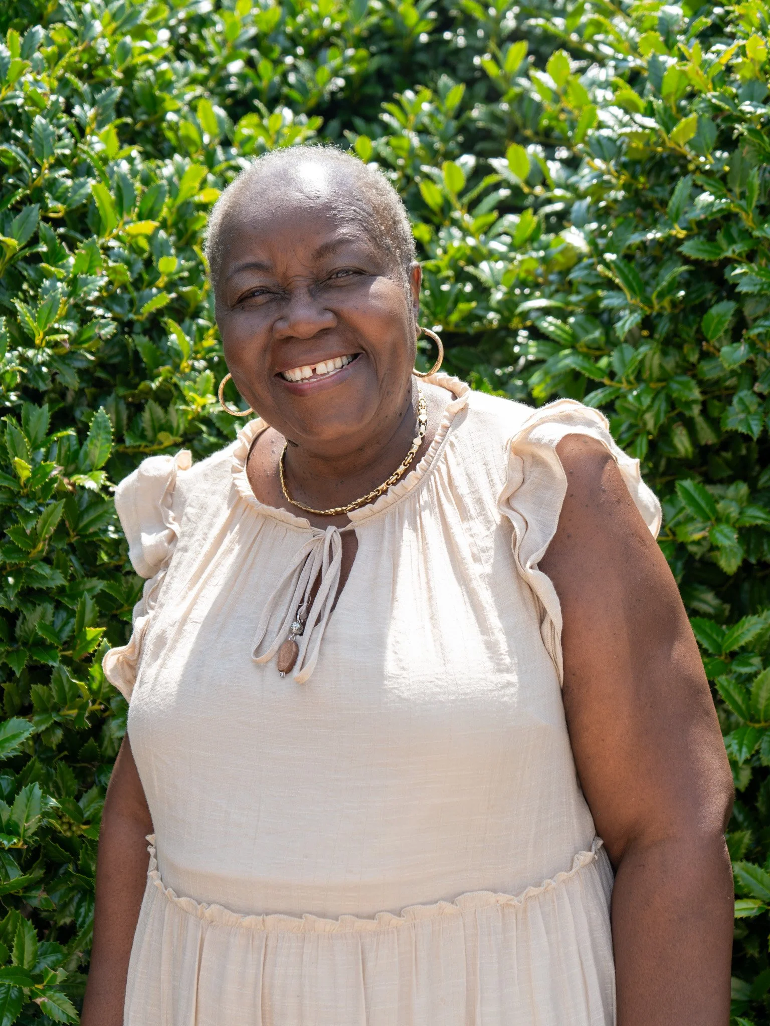 A smiling elderly woman with short gray hair wearing a multicolored sweater and hoop earrings, sitting in a room with yellow walls and wooden decor.