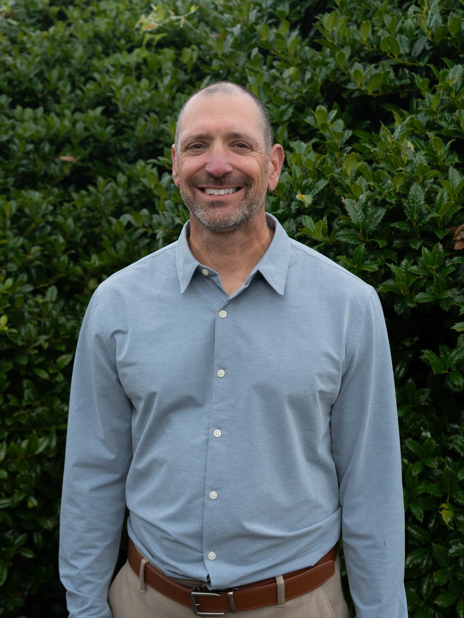 A smiling man with a beard and short hair wearing a black button-up shirt standing against a plain white wall.