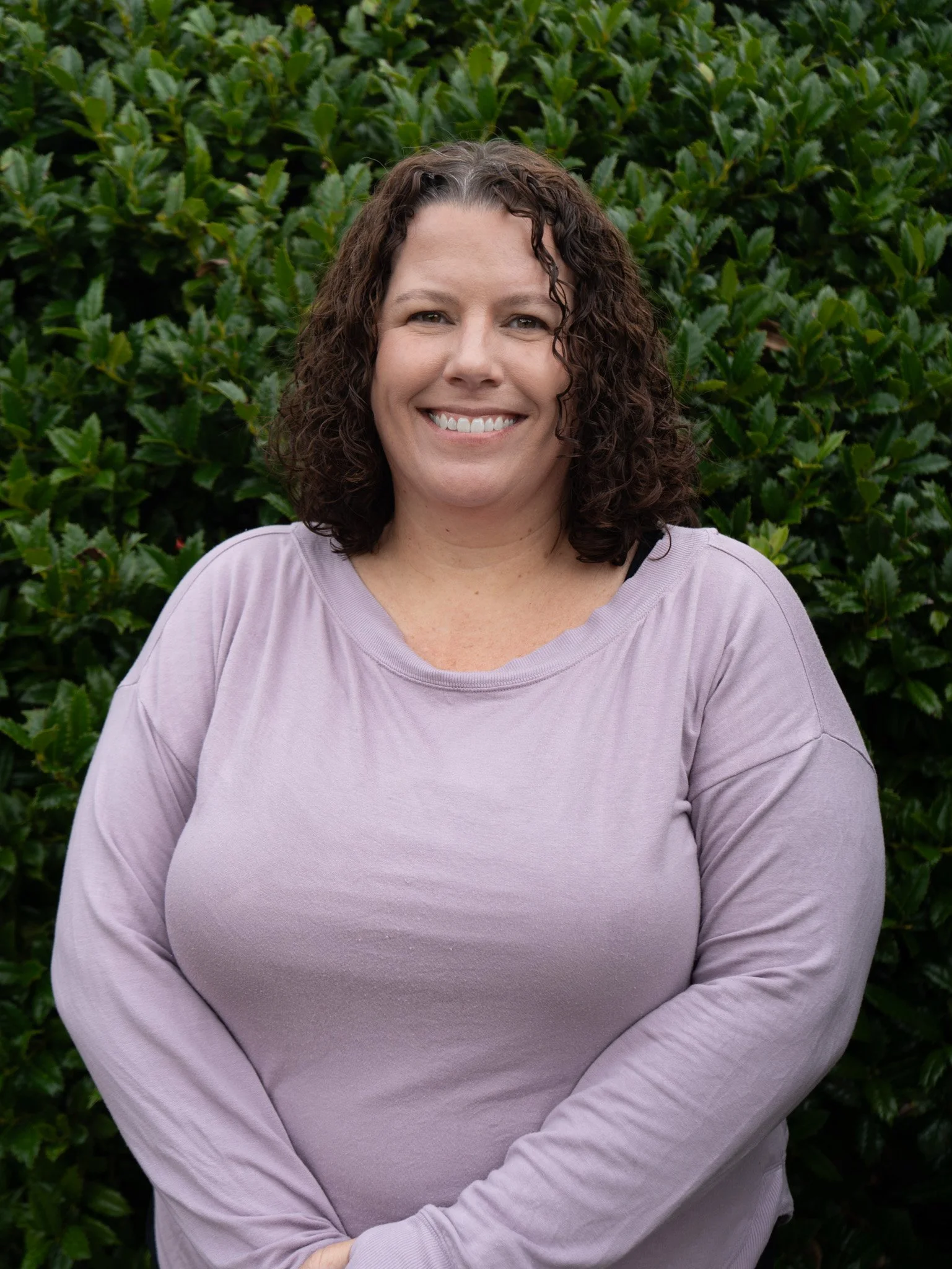 Close-up of a smiling person wearing a purple sweatshirt, with dark hair pulled back, standing in front of a decorative wrought iron wall hanging.