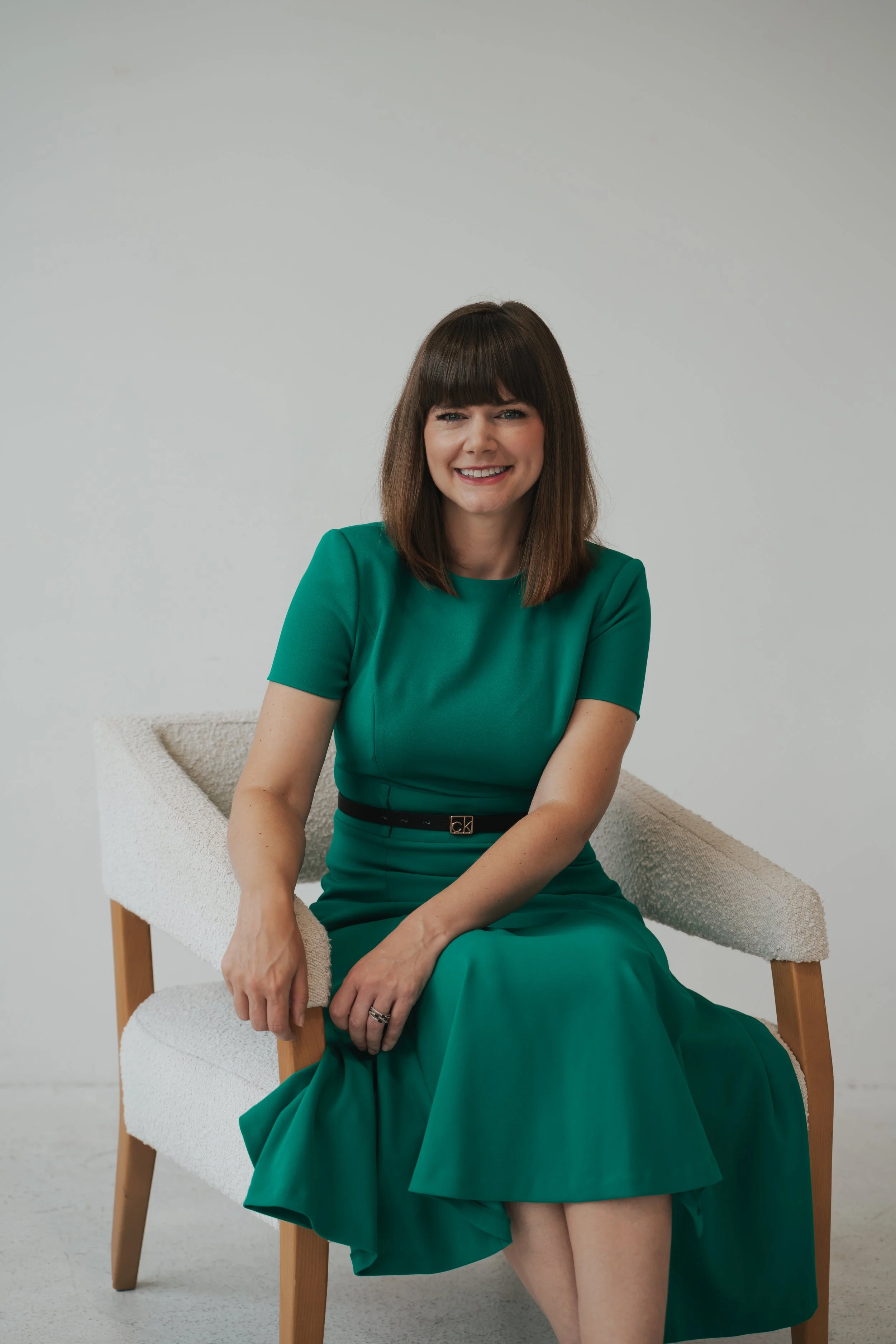 A woman with shoulder-length brown hair and bangs, wearing a green dress, sitting on a beige chair with wooden legs against a plain white background.