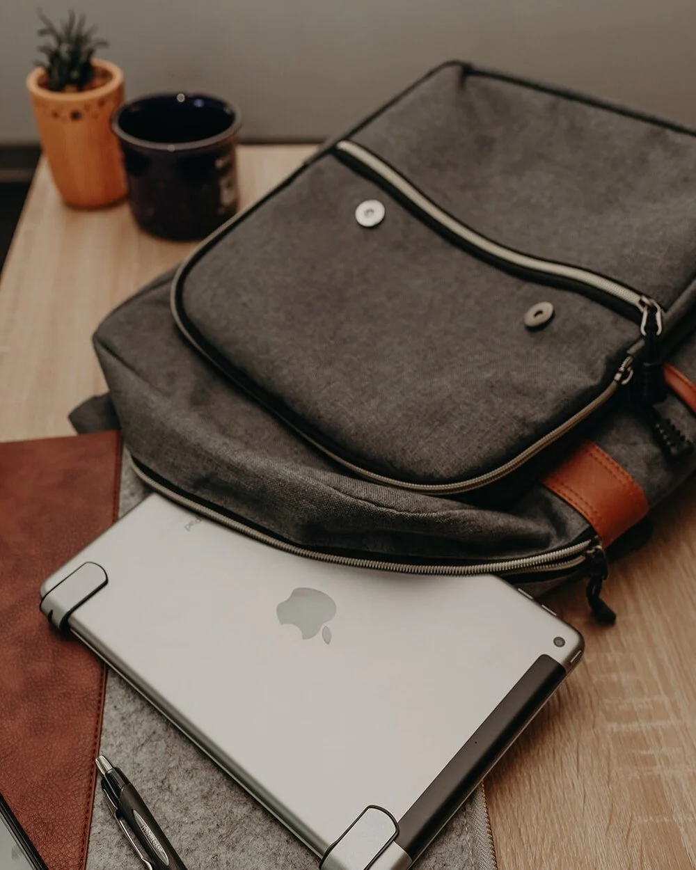 A wooden table with a gray shoulder bag, a silver iPad, a black pen, a small potted cactus, and a black cup.