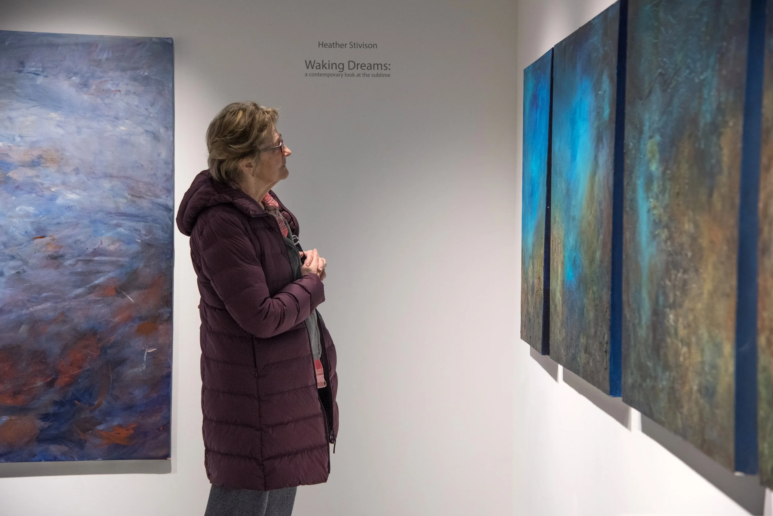 A woman with glasses and a maroon jacket viewing colorful abstract paintings in an art gallery. The gallery wall behind her displays the paintings, and there is text on the wall that reads 'Heather Stivison, Waking Dreams: a contemporary look at the sublime.'
